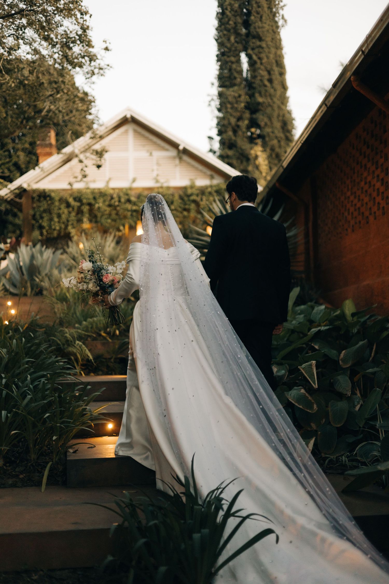 Bride in pearl-adorned veil and gown walks with groom through lush garden at dusk.