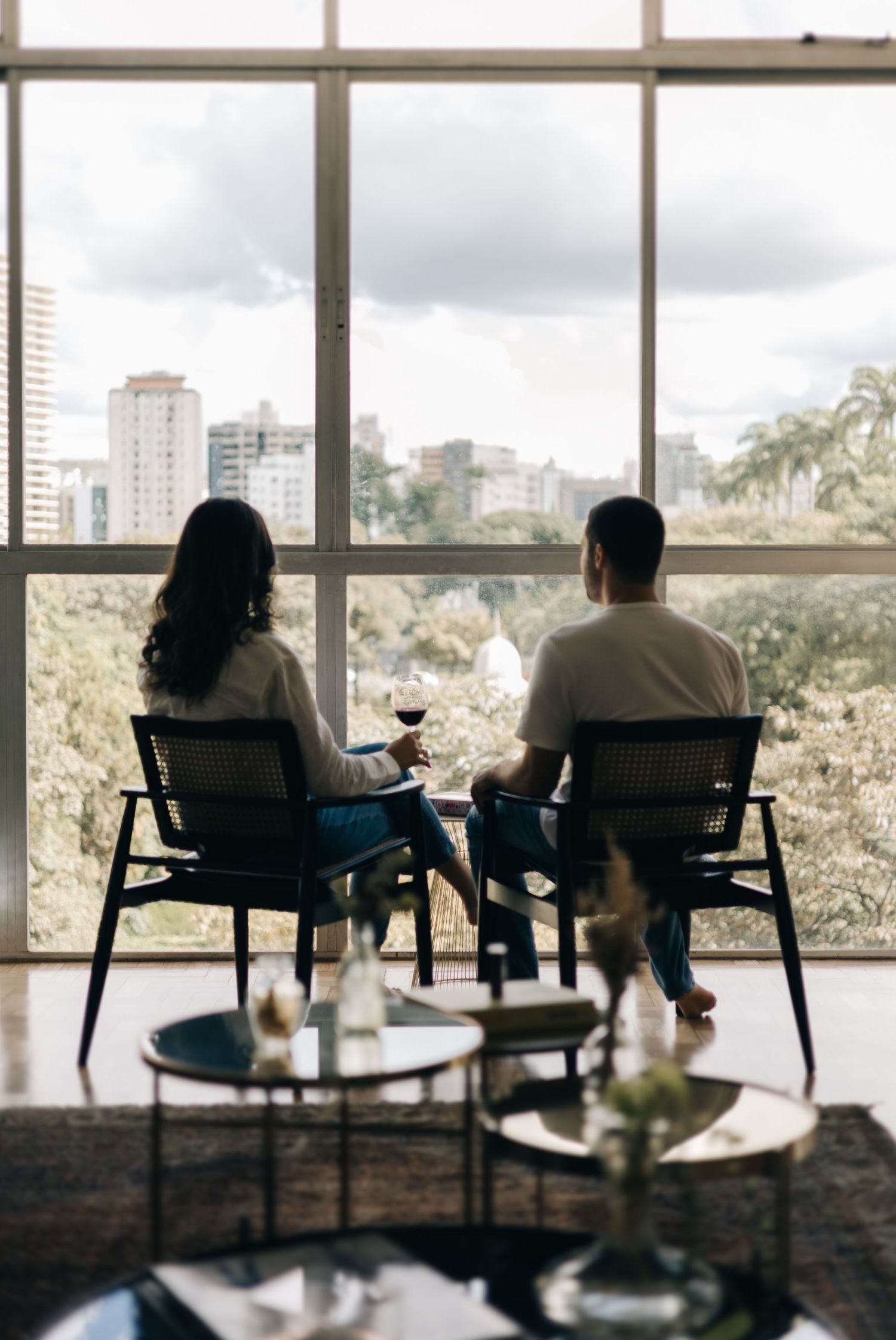 Two people enjoy wine while sitting in modern chairs by a large window overlooking a city skyline and greenery.
