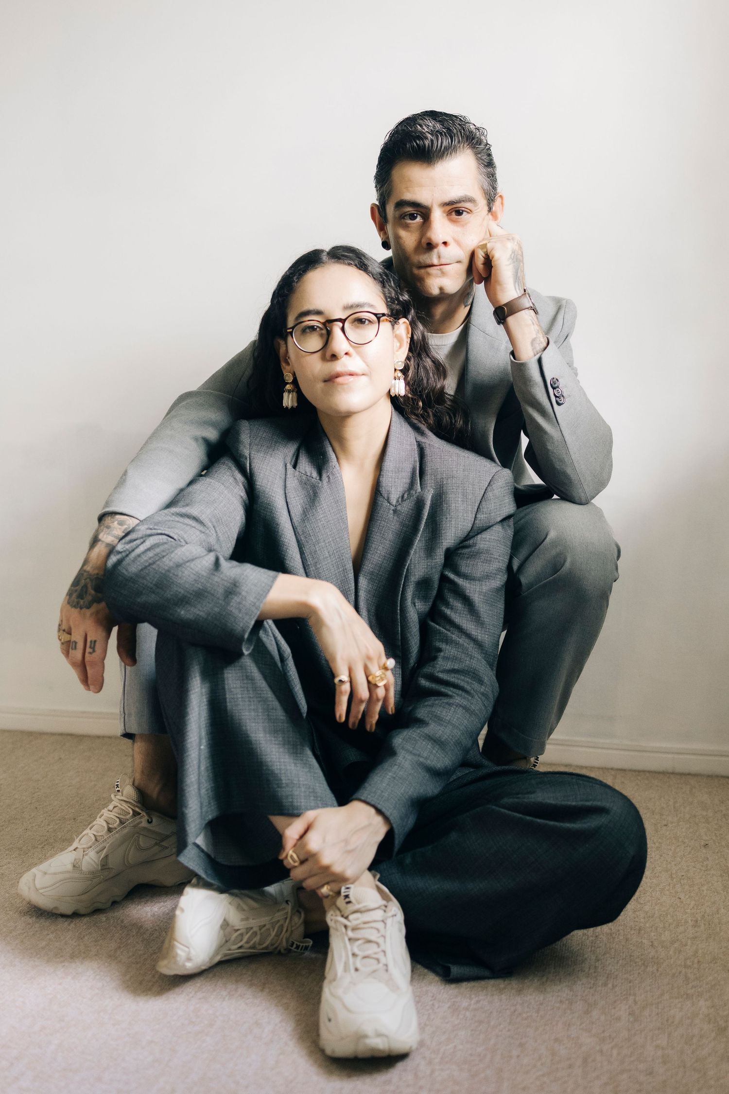 Two people in gray business suits sit casually on the floor in a minimalist studio setting.