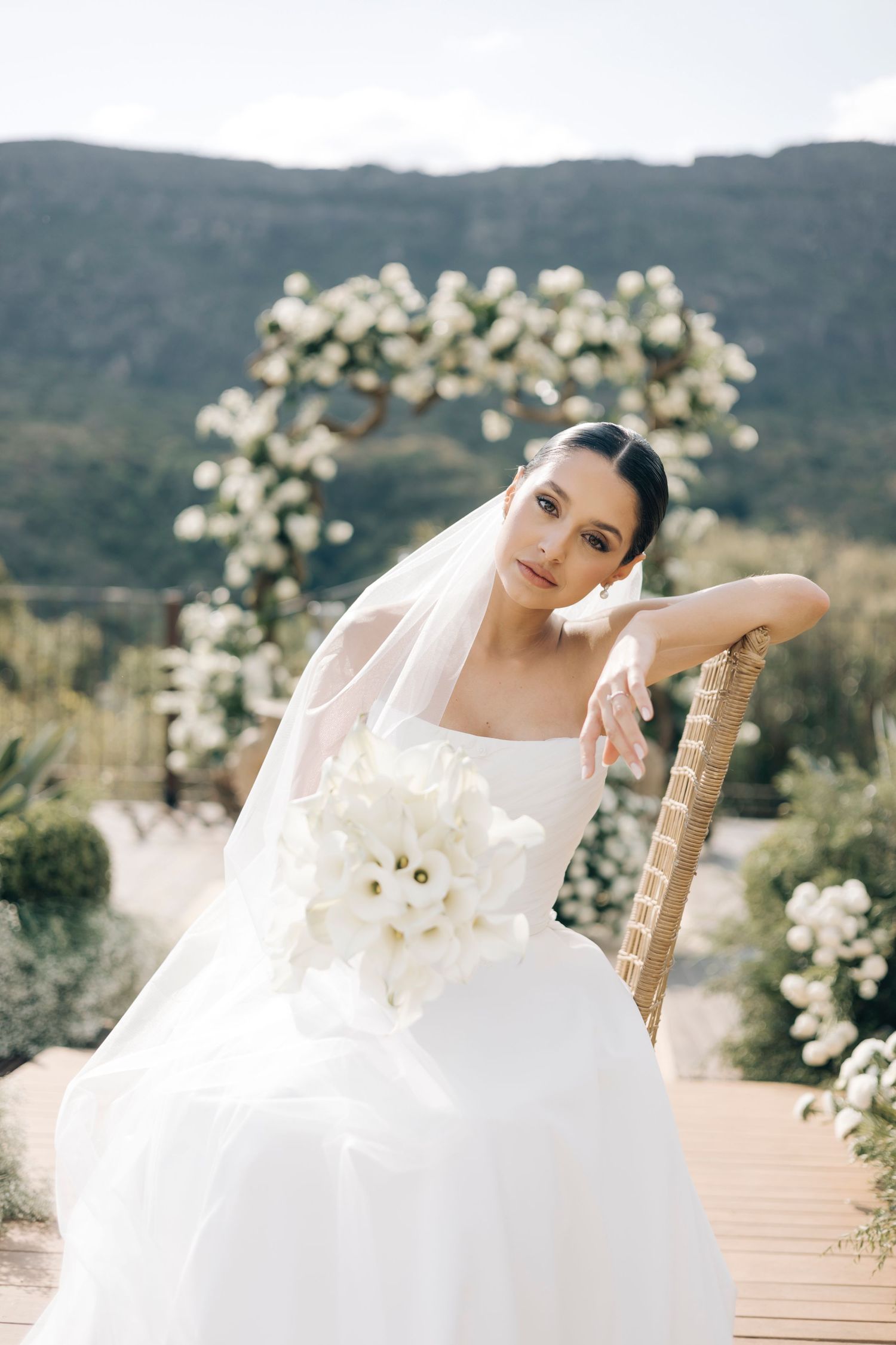 A bride in a flowing white gown poses elegantly outdoors with white floral arrangements and mountains in the background.