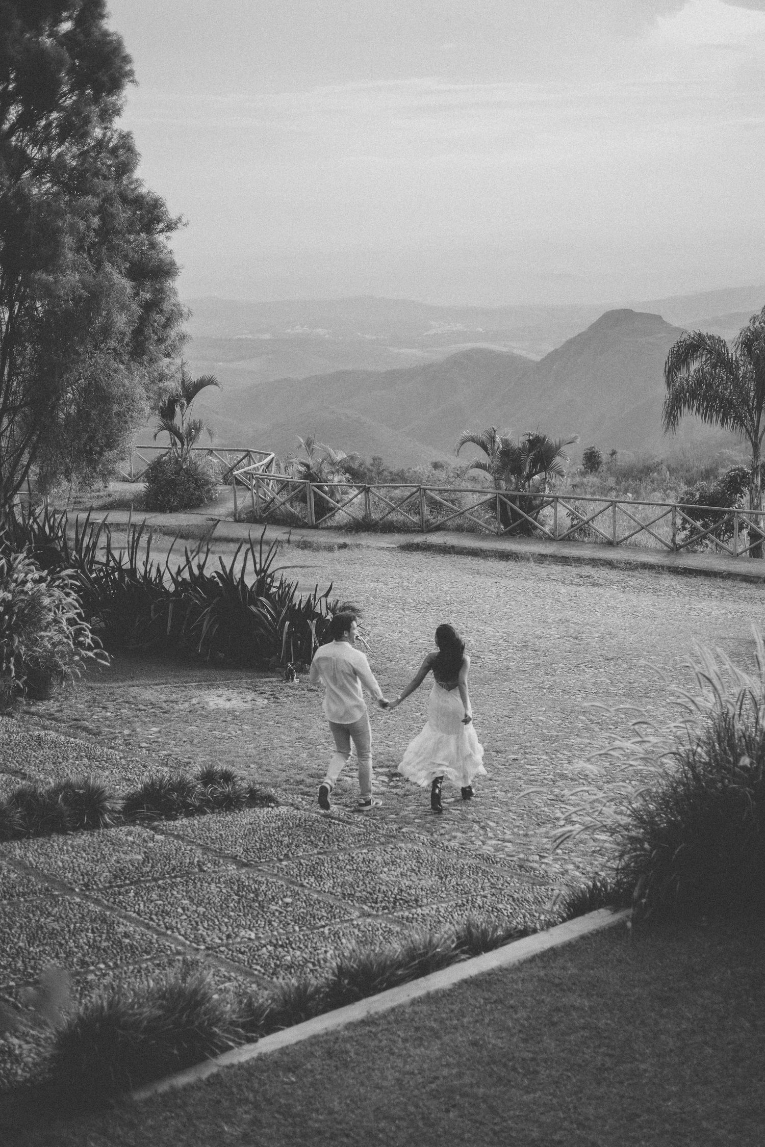 Black and white photo of two people walking together on a scenic mountain path with a view of distant hills and palm trees.