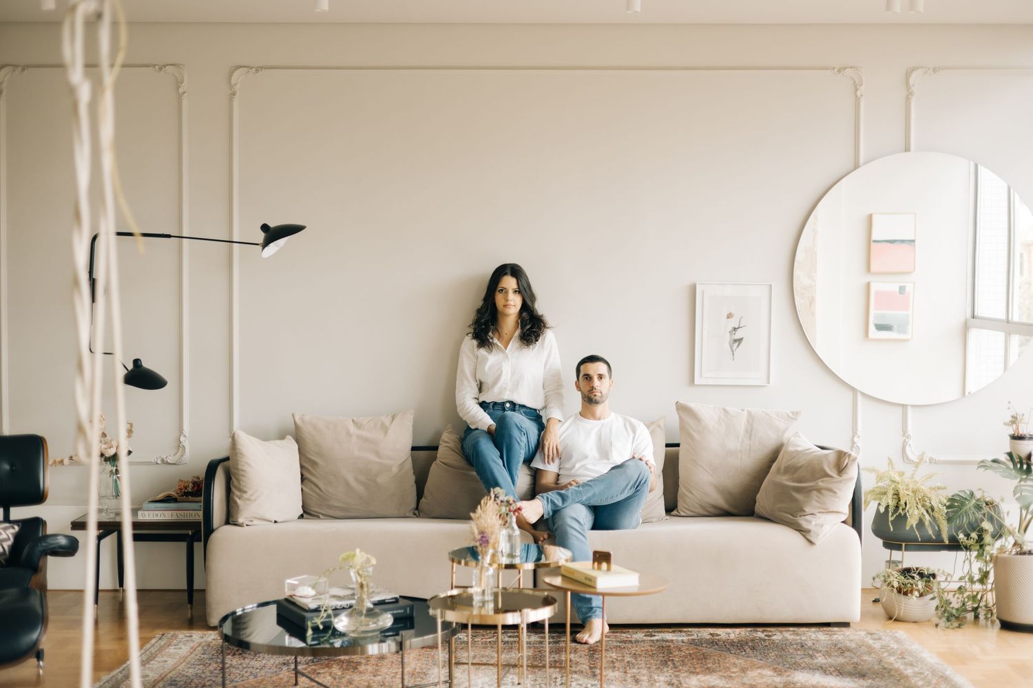 A couple lounges casually on a light gray couch in a minimalist living room with natural lighting and plants.