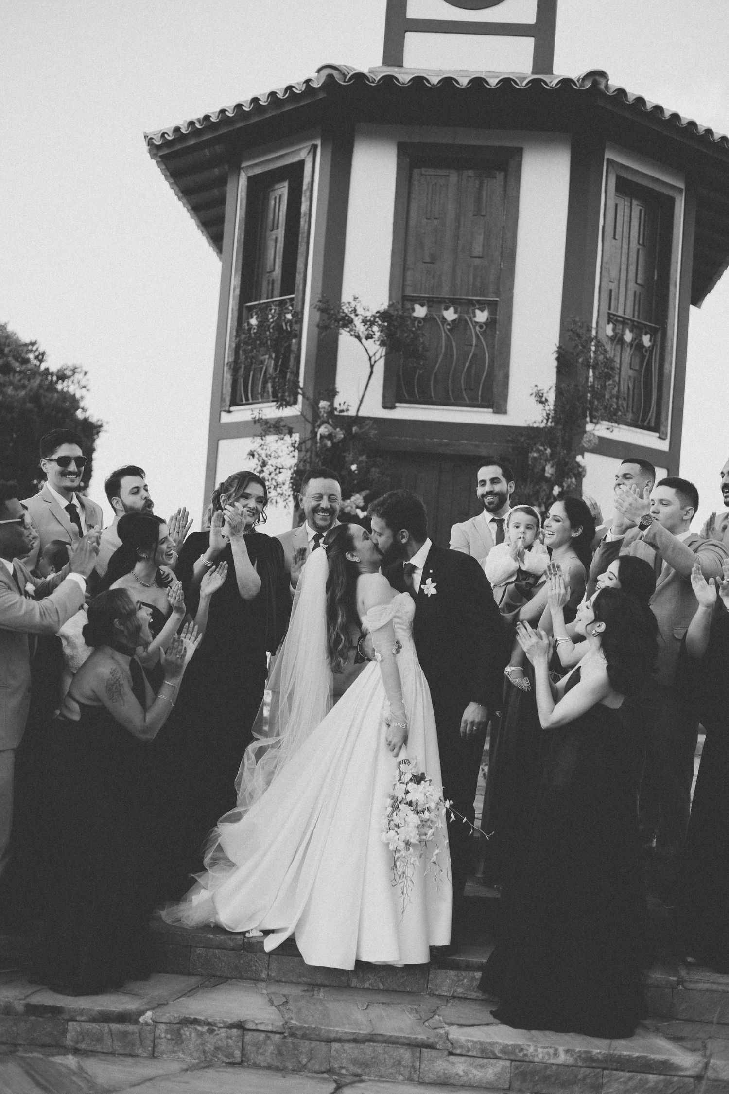 A black and white wedding photo shows guests celebrating around a newlywed couple near a tower entrance.