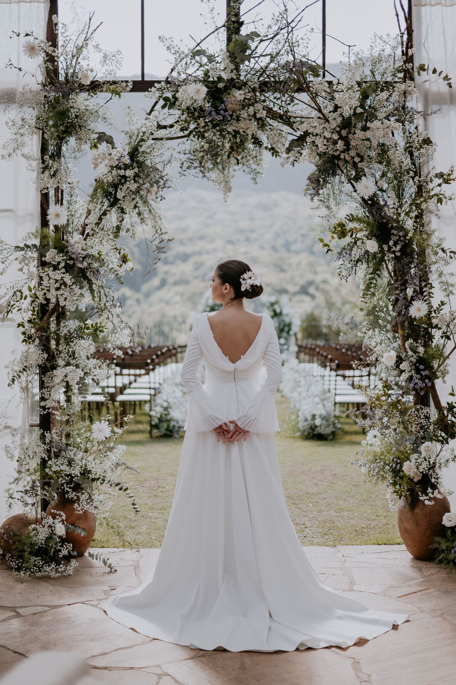 Bride in flowing white dress standing under floral arch with mountain backdrop.