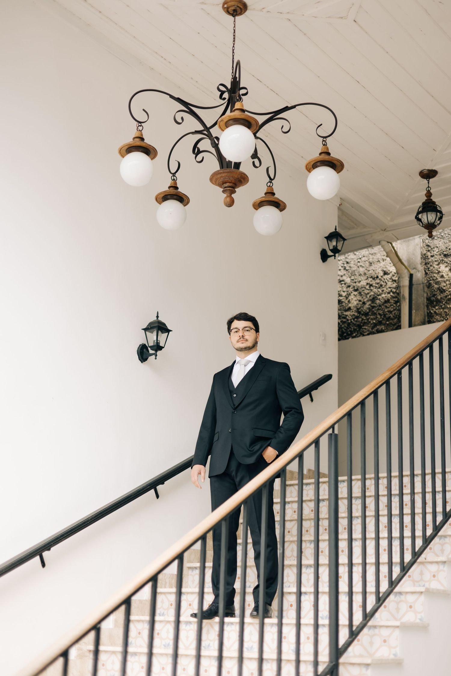 Groom in classic black suit poses on staircase under vintage chandelier in bright interior.
