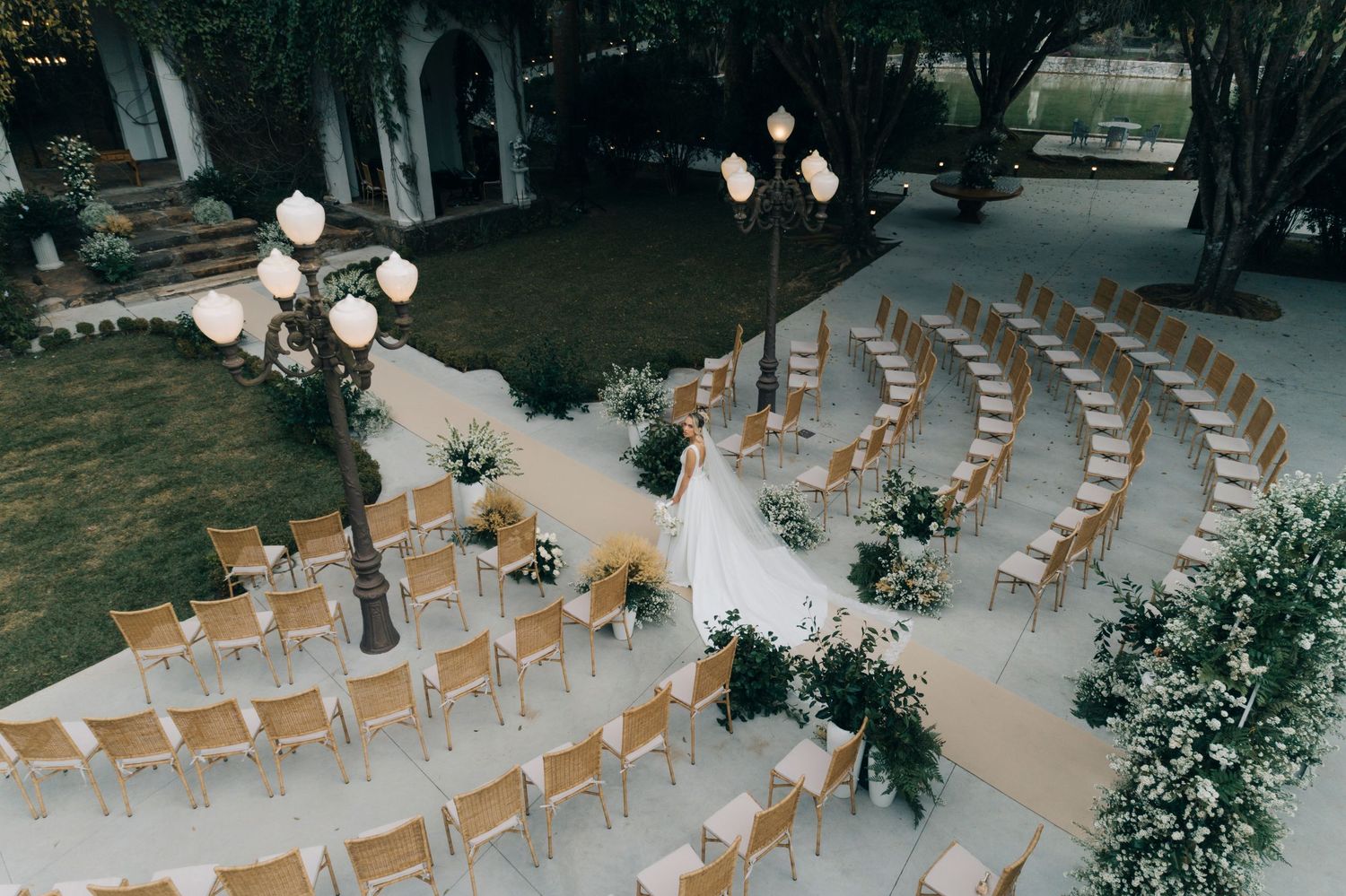 White wooden chairs arranged in rows outdoors for a garden wedding ceremony under string lights and flowering trees.