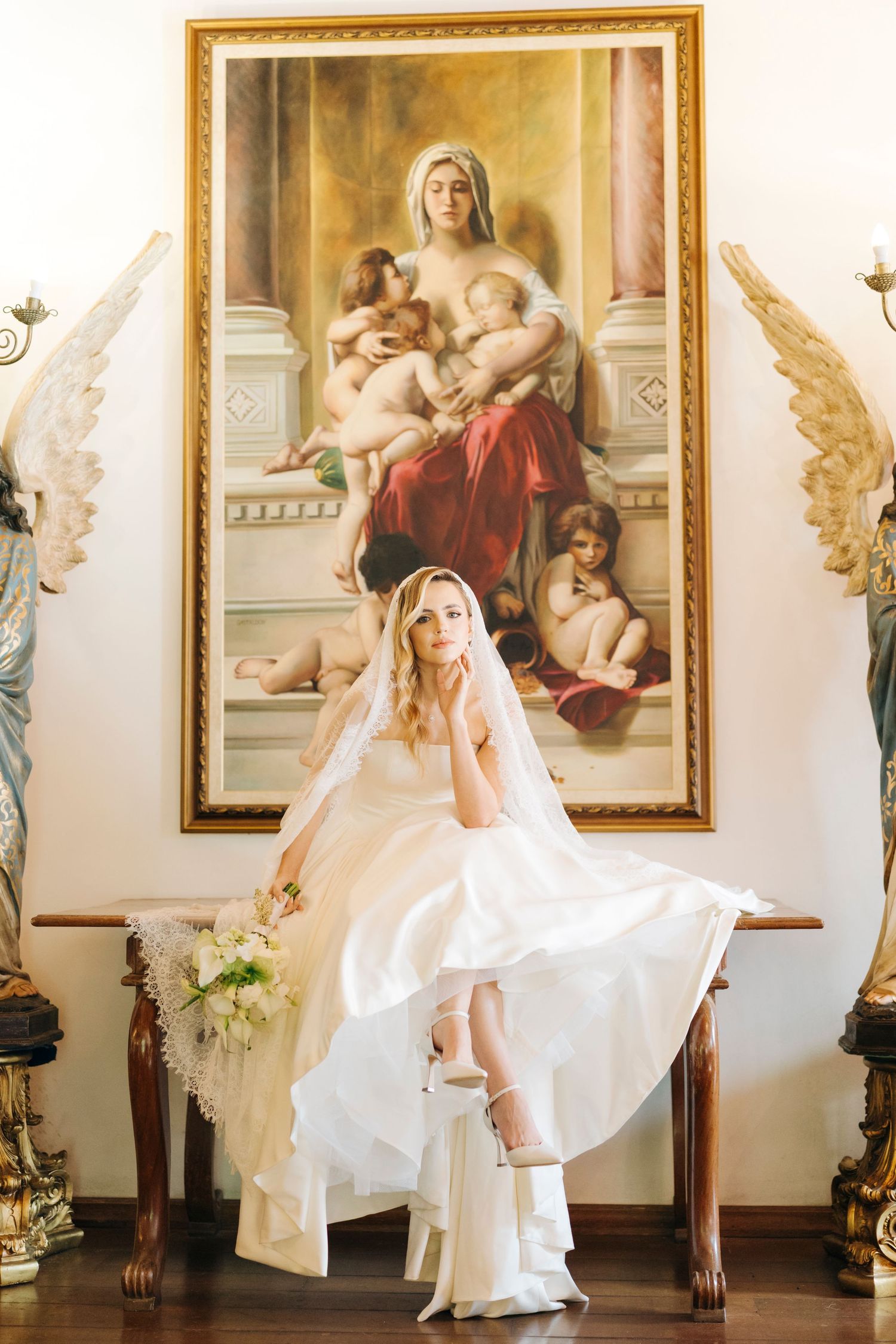 Bride in white gown and veil sits before a classical religious painting flanked by angel statues holding a bouquet.