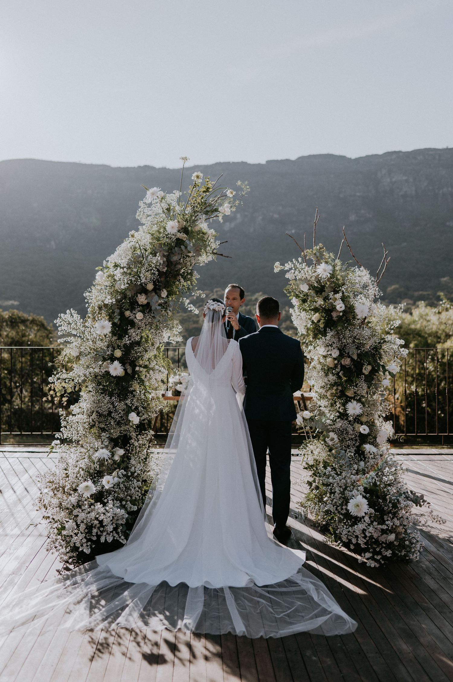 Wedding ceremony under white floral arch with mountain vista backdrop.