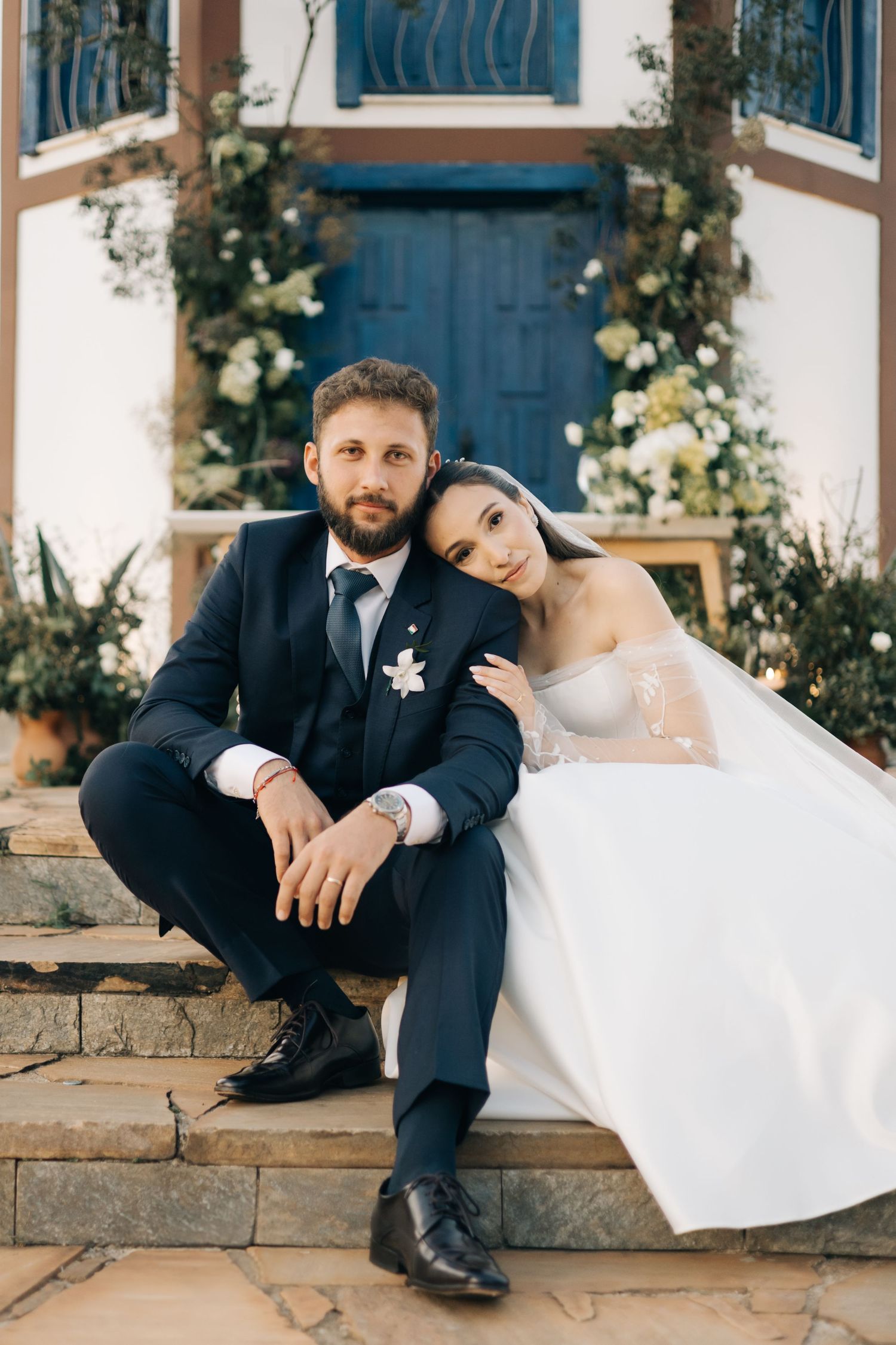 A couple sits on stone steps in wedding attire with a beautiful blue door and floral decorations behind them.