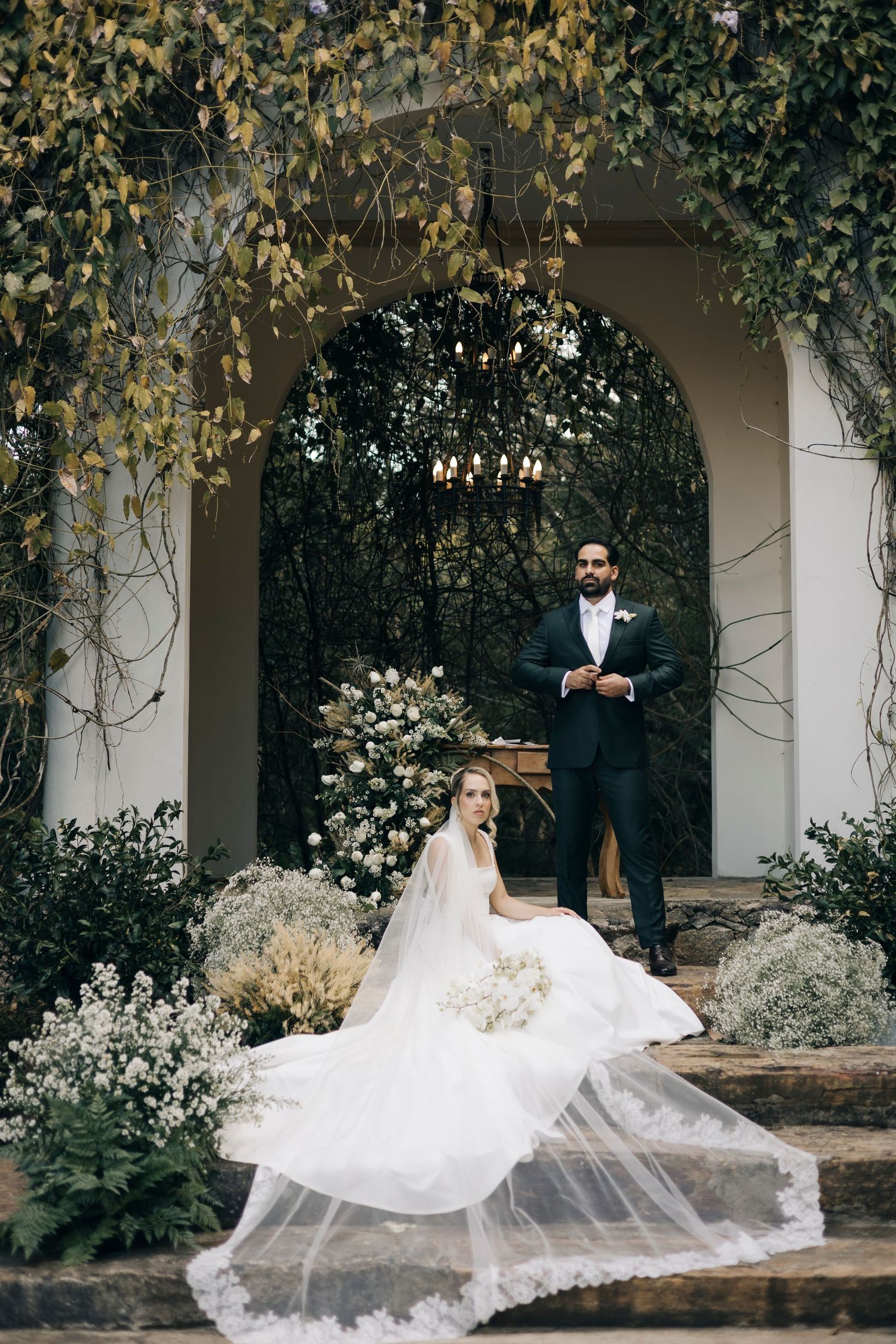 A couple poses on stone steps under an archway with flowing veil and floral decorations in an elegant garden setting.