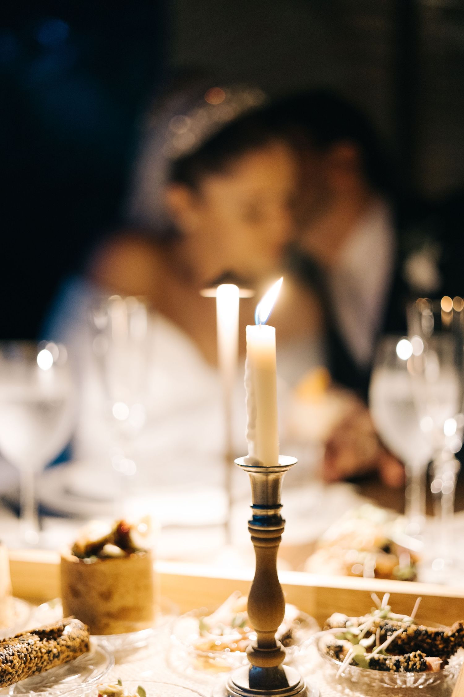 A romantic candlelit dinner with a couple kissing in the blurred background, elegant table setting with wine glasses and food.