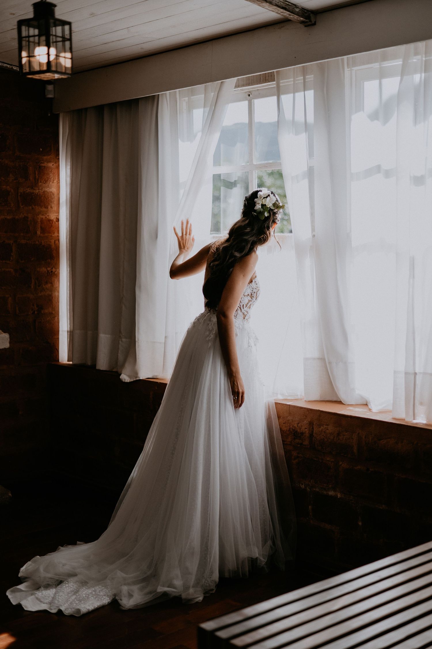 Bride in flowing white gown peeking through sheer curtains in moody interior lighting.