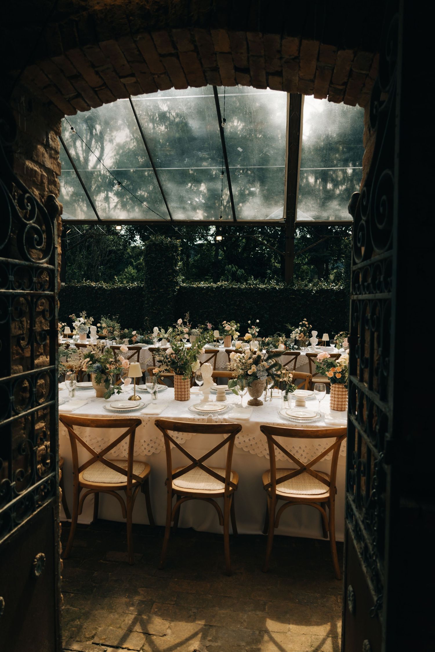 Romantic wedding reception setup viewed through rustic brick archway with vintage wooden chairs.