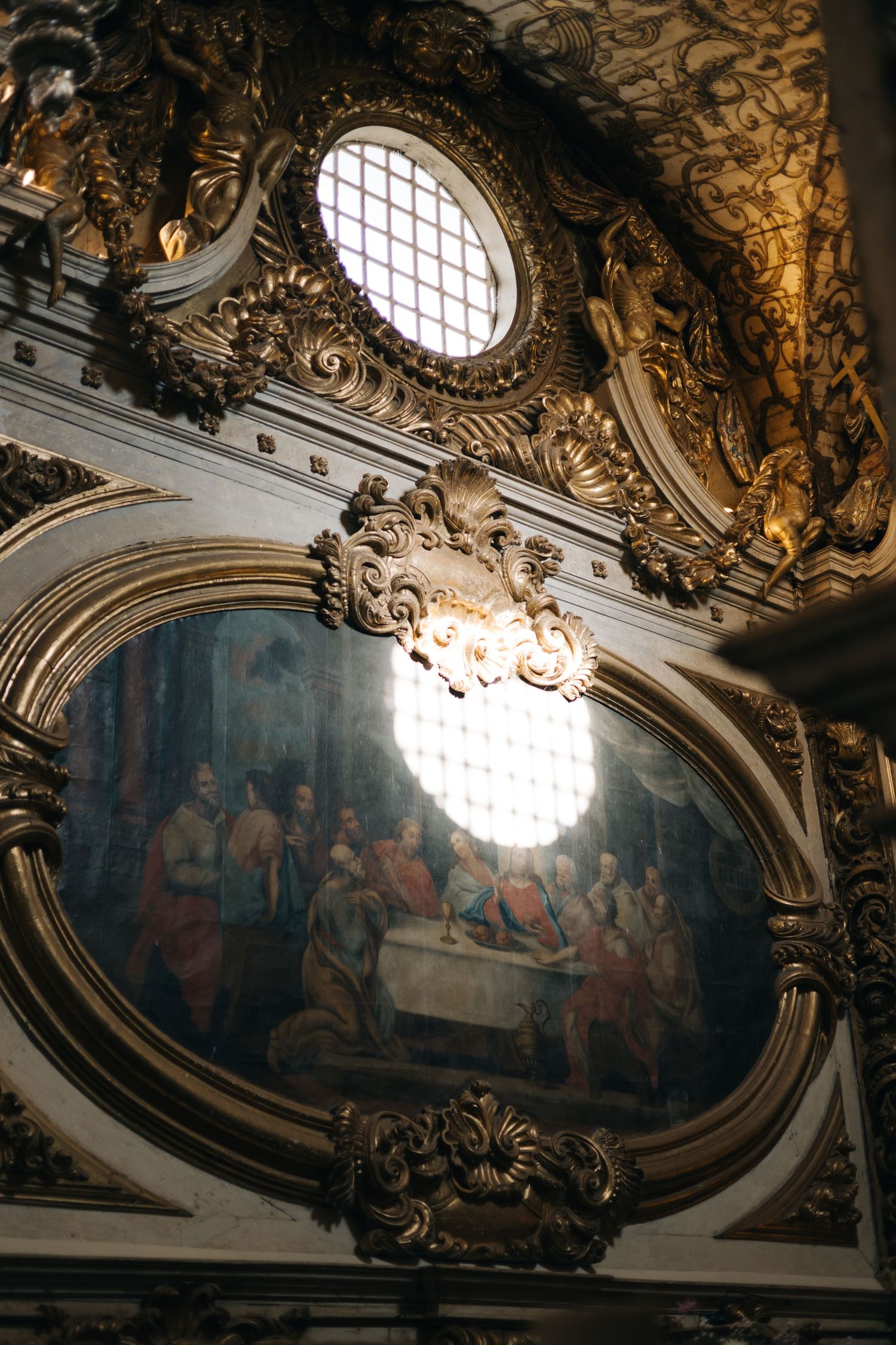 Ornate baroque ceiling with gilded details and a round window illuminating a religious fresco painting.