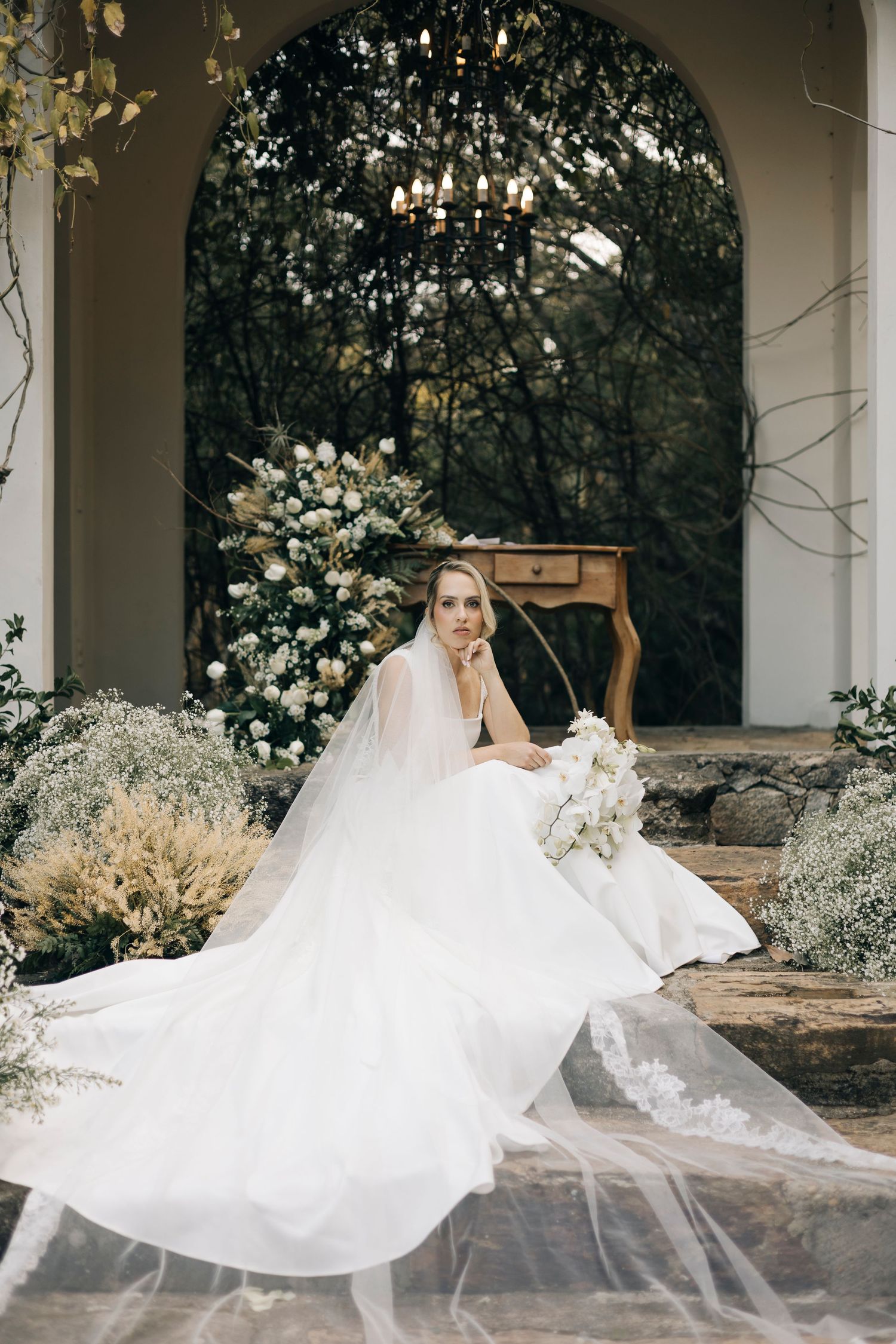 A flowing white wedding dress and veil billow dramatically in an elegant garden archway setting.