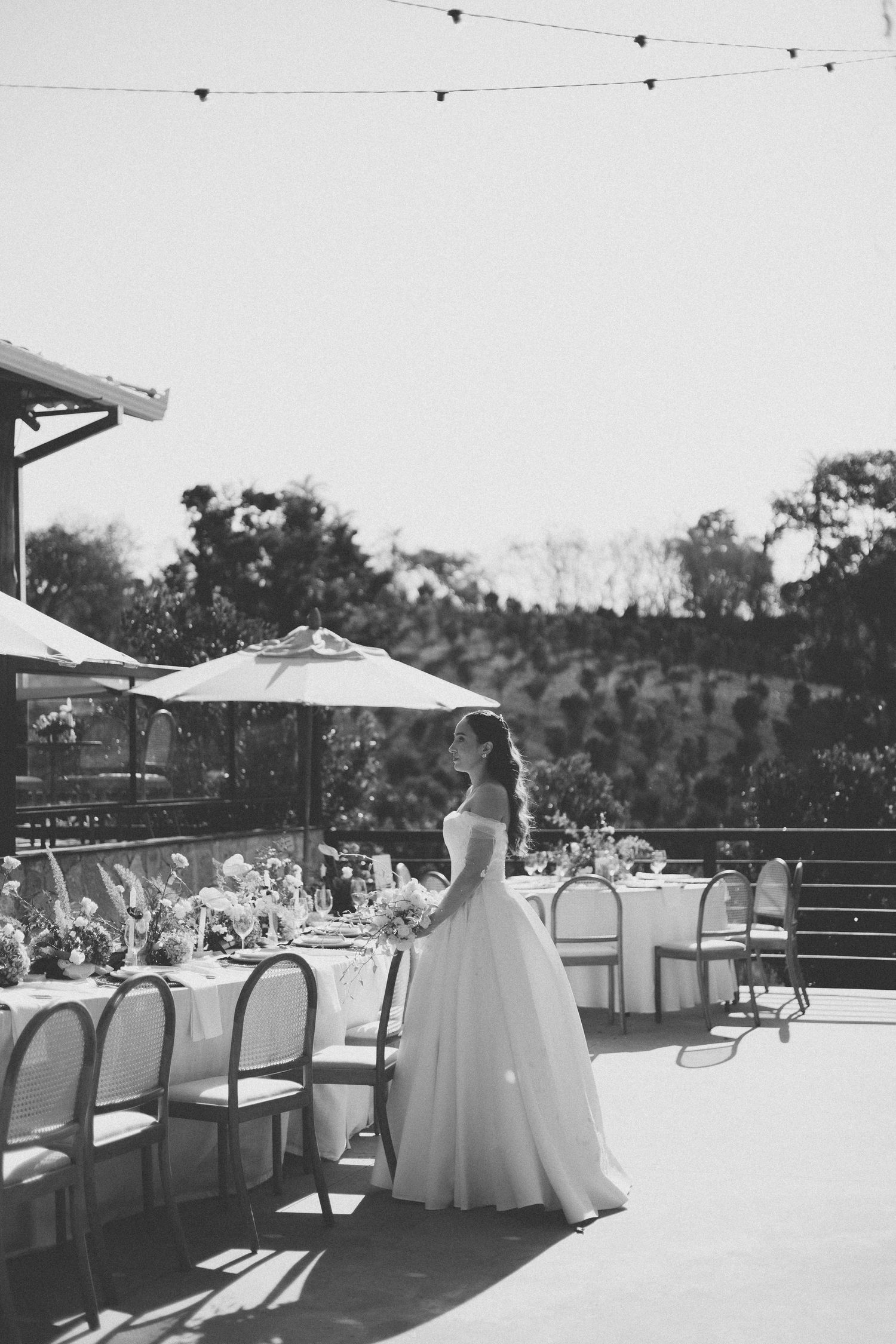 A bride in a flowing white gown stands on a sunlit outdoor terrace with string lights and empty chairs.