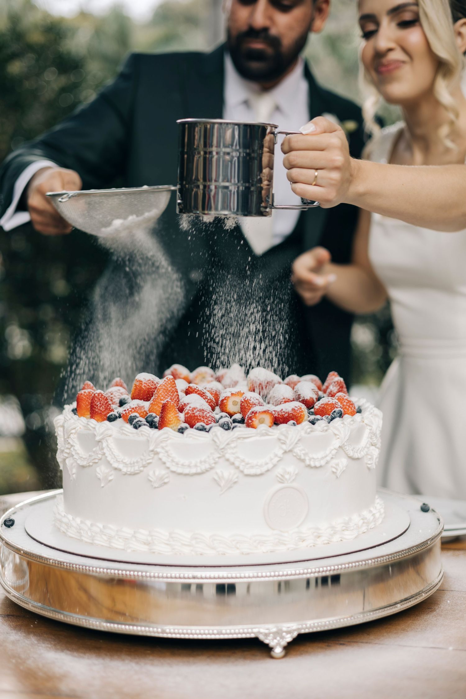 A white wedding cake decorated with fresh berries is being dusted with powdered sugar during a wedding celebration.