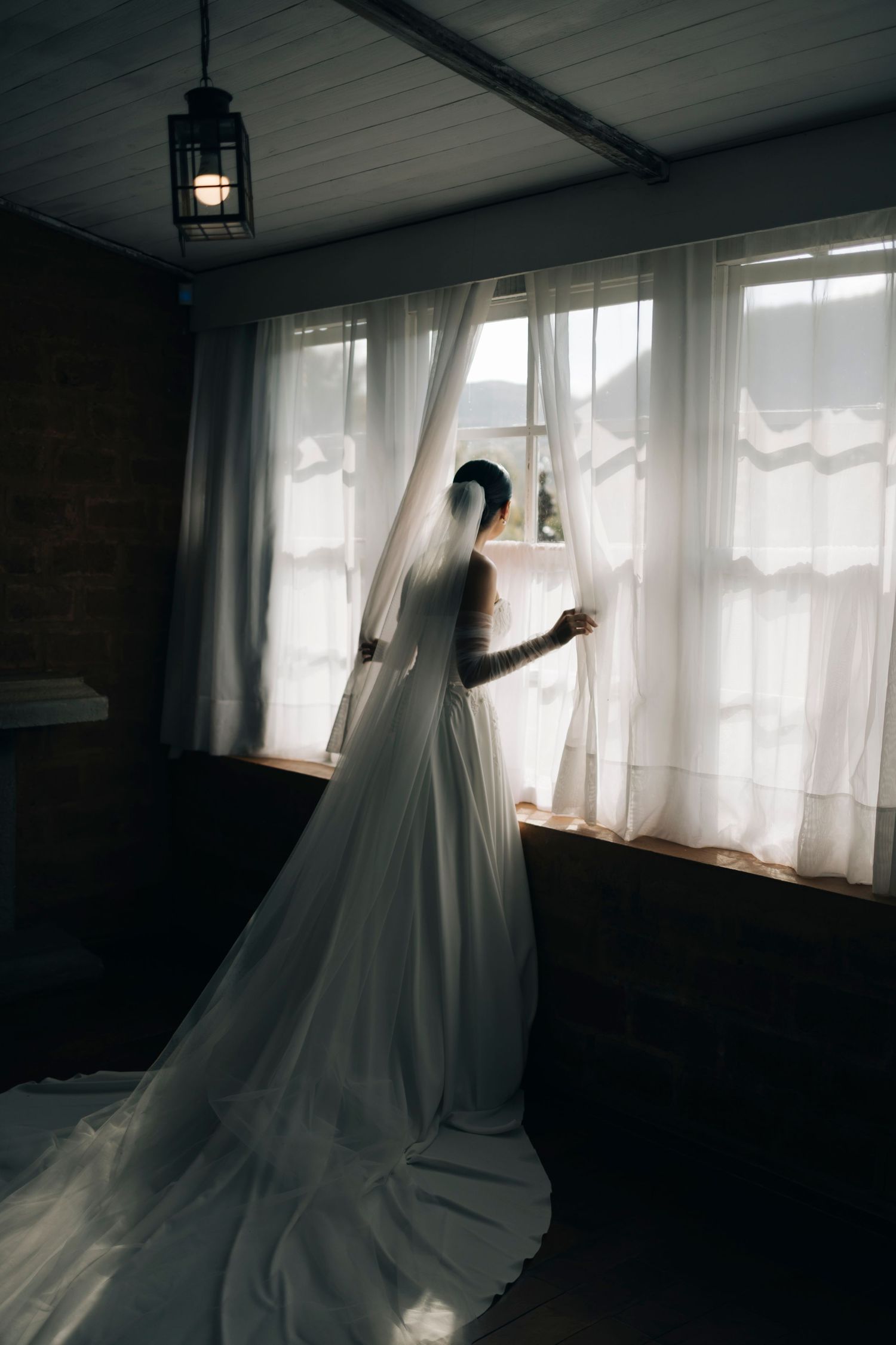 Bride in white wedding gown and veil stands by sunlit window with sheer curtains in rustic room.
