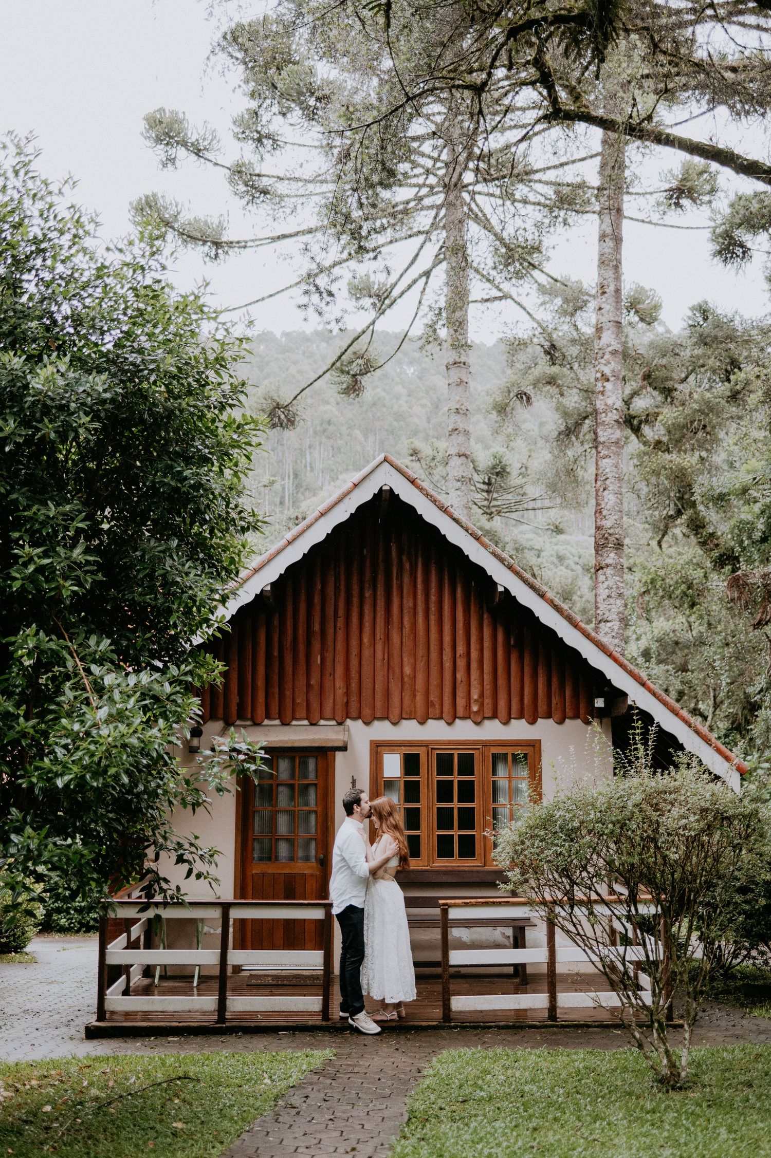 Cozy rustic wooden cabin in the woods with French doors, front deck and tall pine trees surrounding it on a misty day.