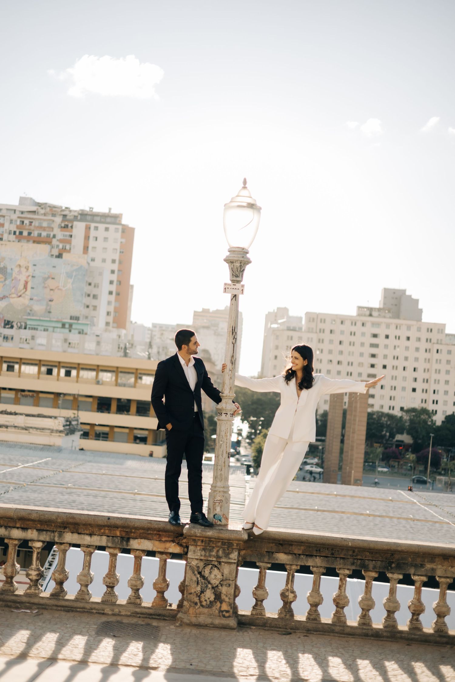 A couple stands on an ornate balcony with vintage lamp post and city buildings in the background on a sunny day.