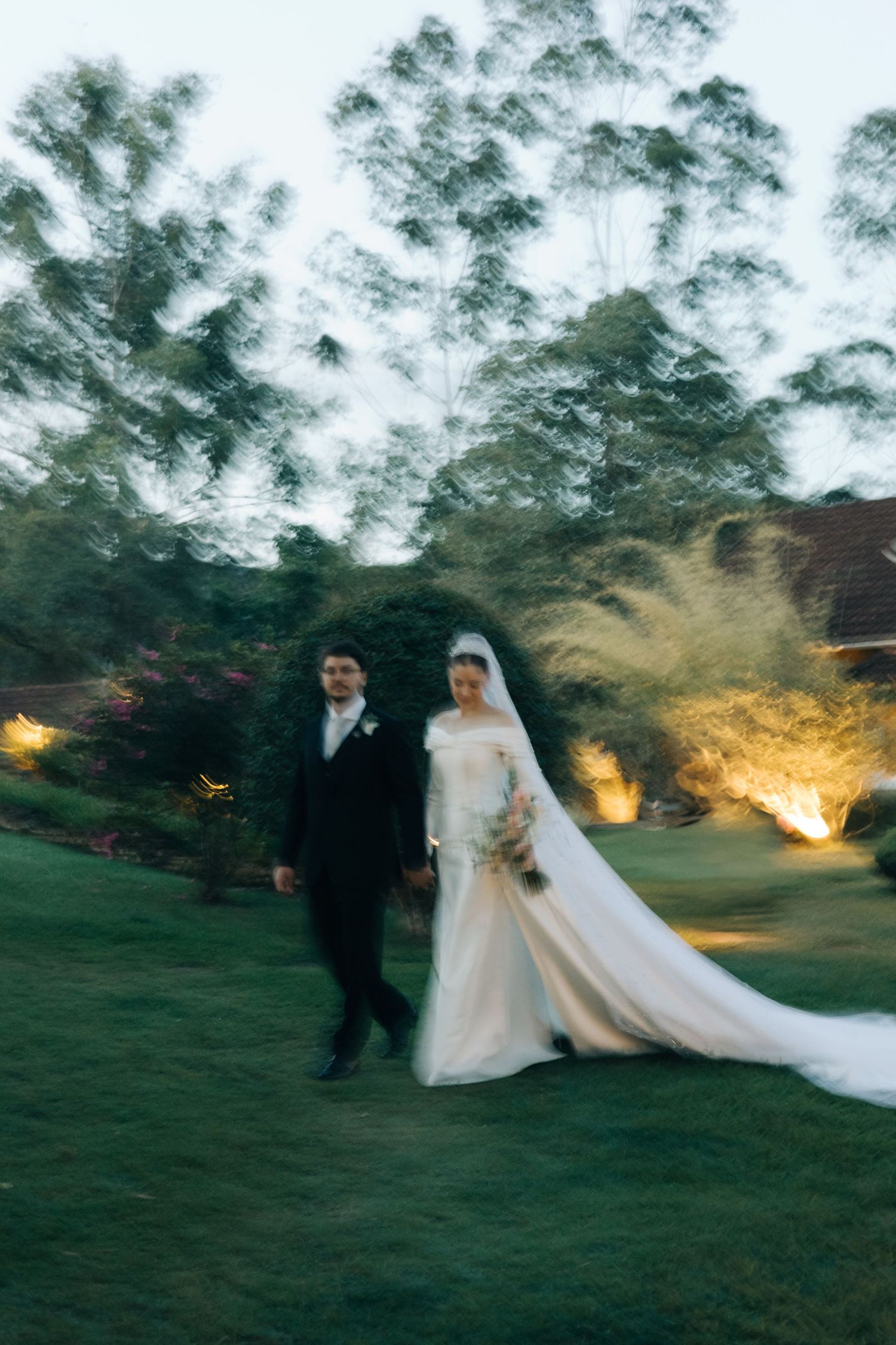Newlyweds stroll through a twilight garden with illuminated trees in the background.