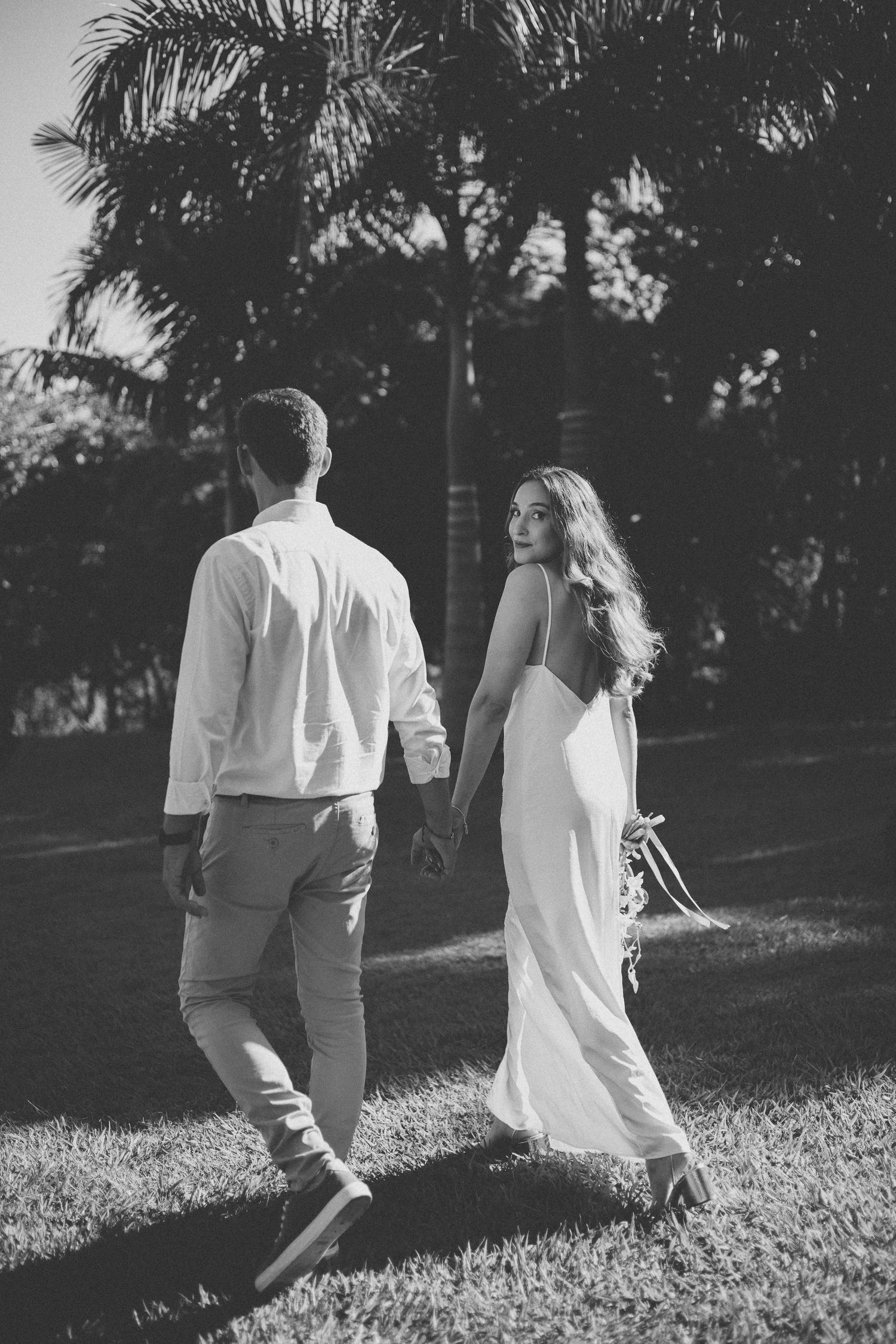 A couple in light-colored attire walks together on grass with palm trees in the background, captured in black and white.