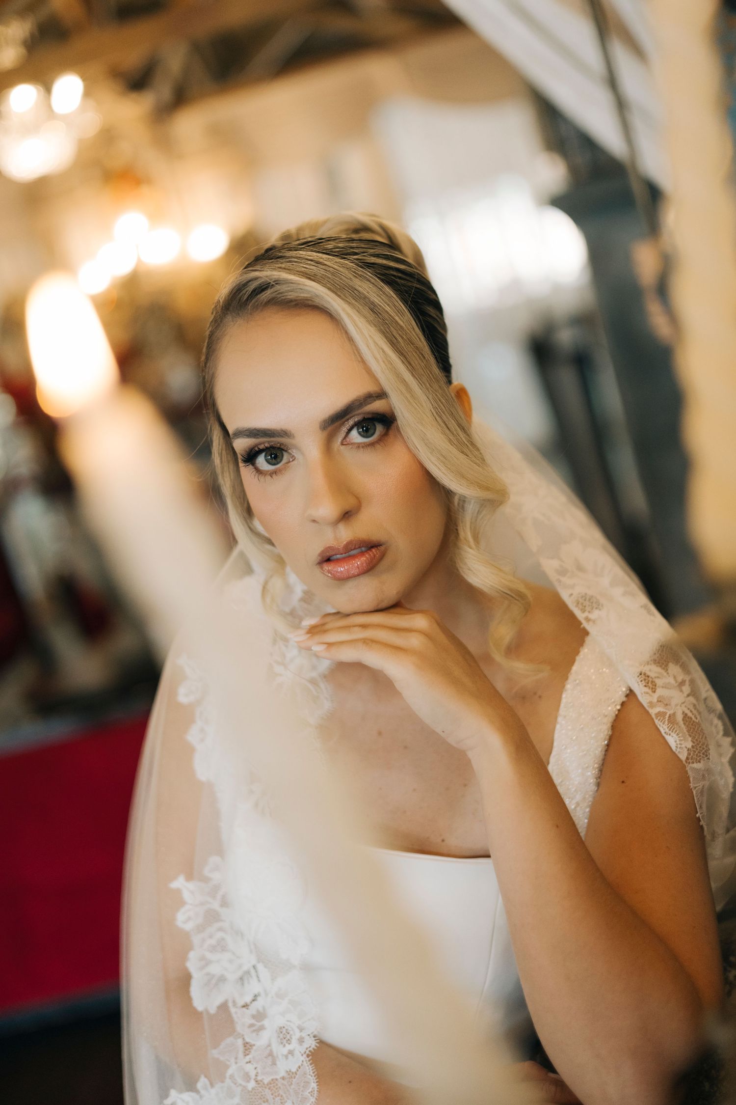 A bride in an elegant white gown and veil poses thoughtfully in a warmly lit indoor setting with bokeh lights in background.