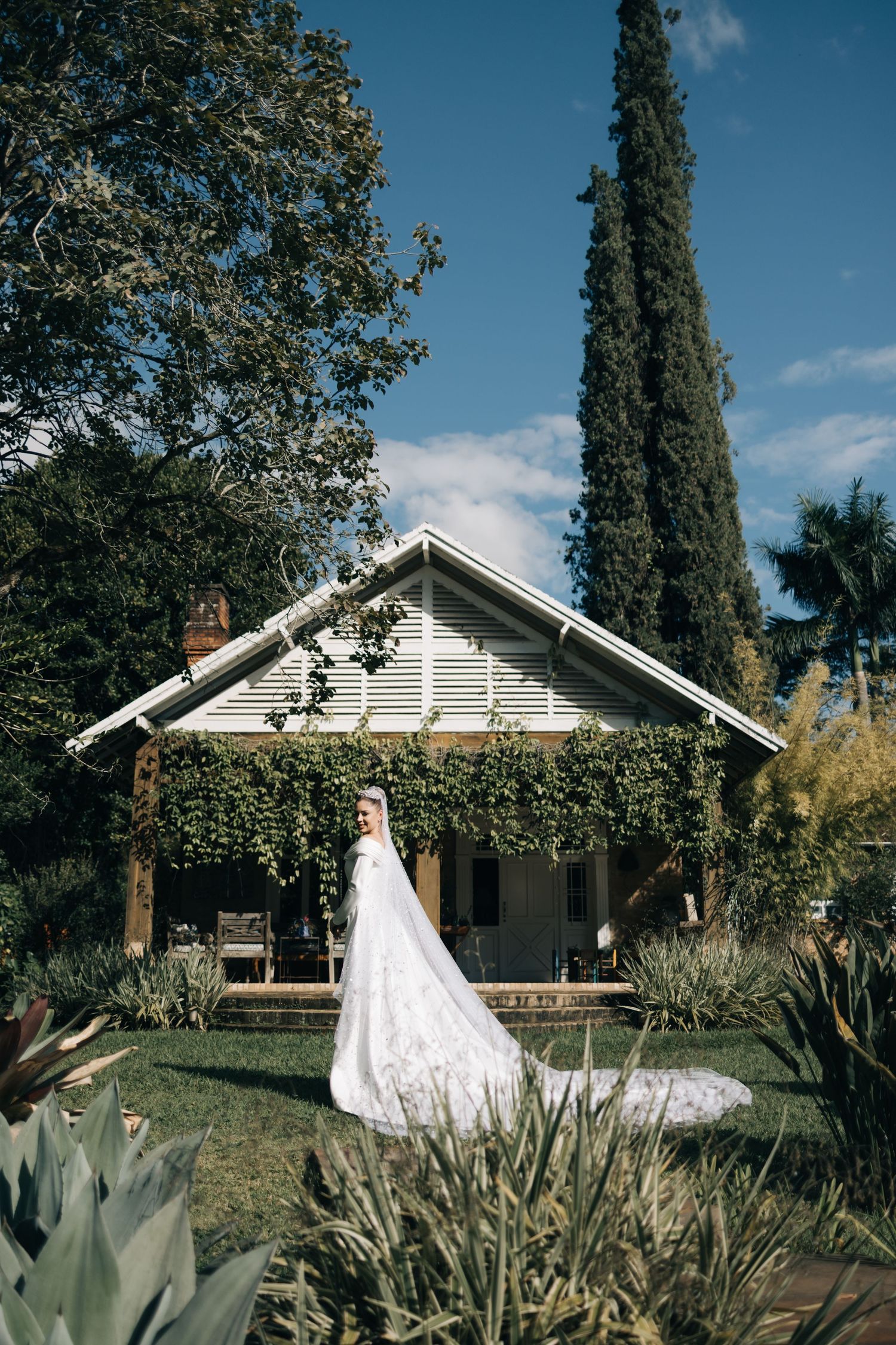 Elegant white wedding dress hanging in front of a charming cottage with cypress trees and lush garden landscaping.