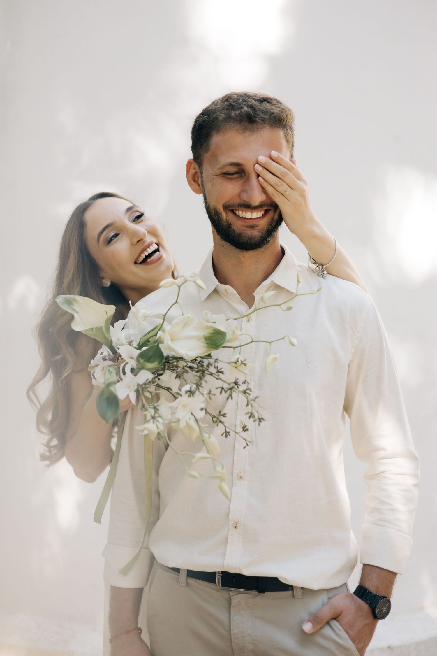 Joyful couple sharing a candid moment while laughing together, with delicate white florals against light background.