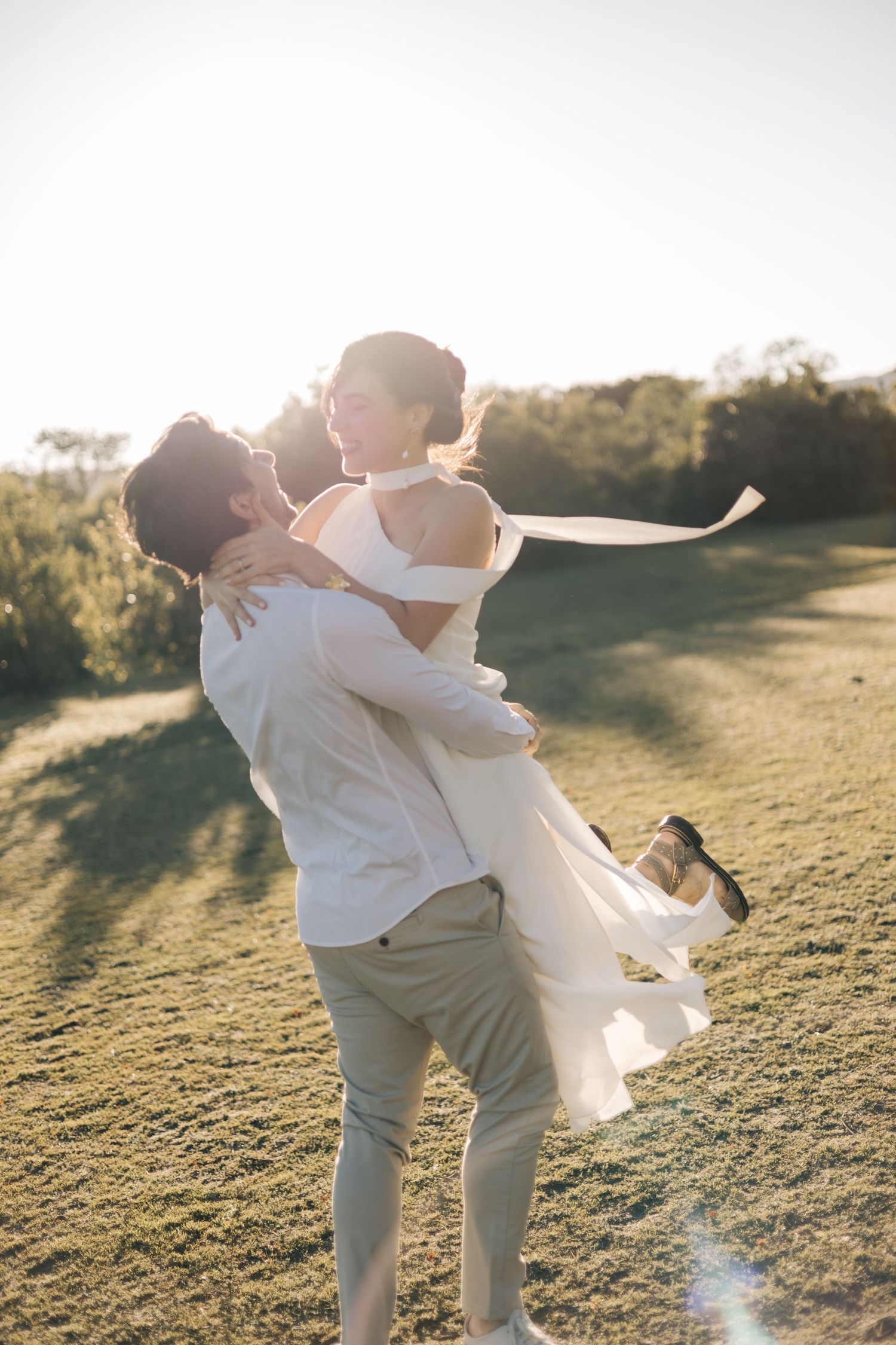 A couple embraces in a romantic twirl against golden sunlight on a grassy hillside during sunset.