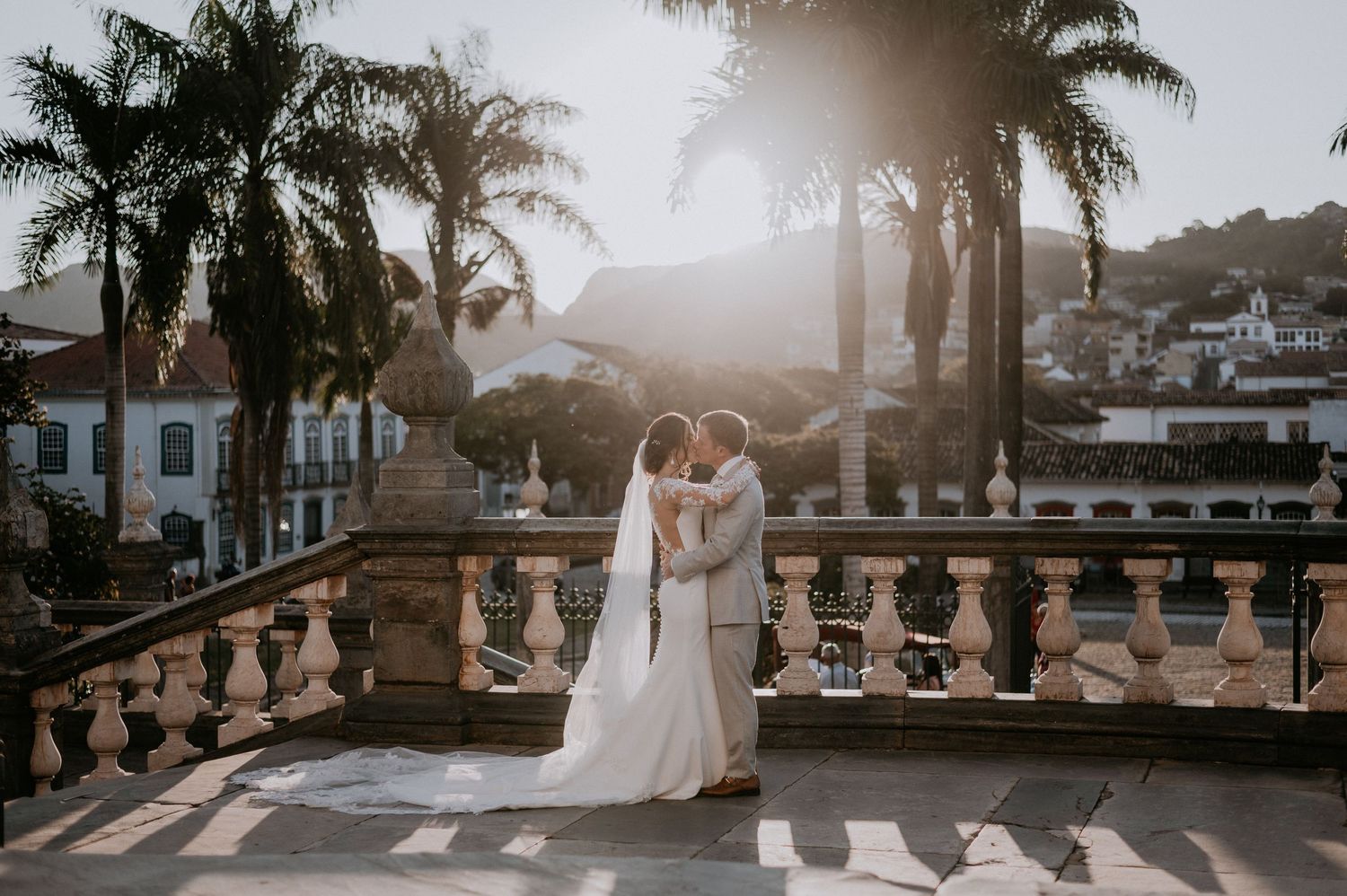 Newlyweds embrace on stone balcony with palm trees during sunset.