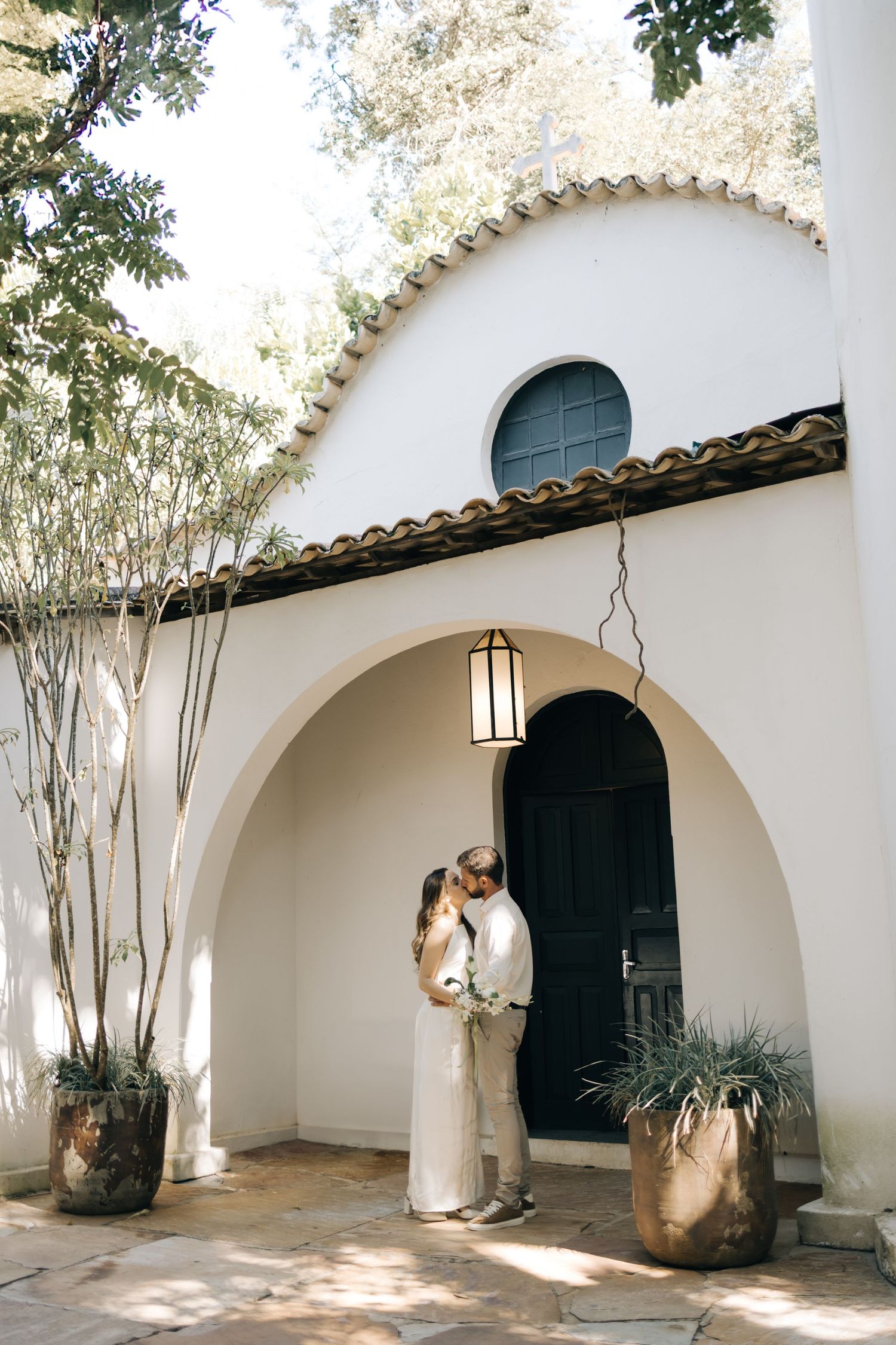 A romantic couple embraces under a Mediterranean-style white archway with a hanging lantern and potted plants.