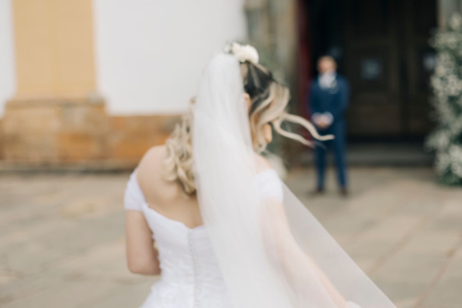 Bride in white wedding dress and veil walks toward groom in blue suit outside a church.