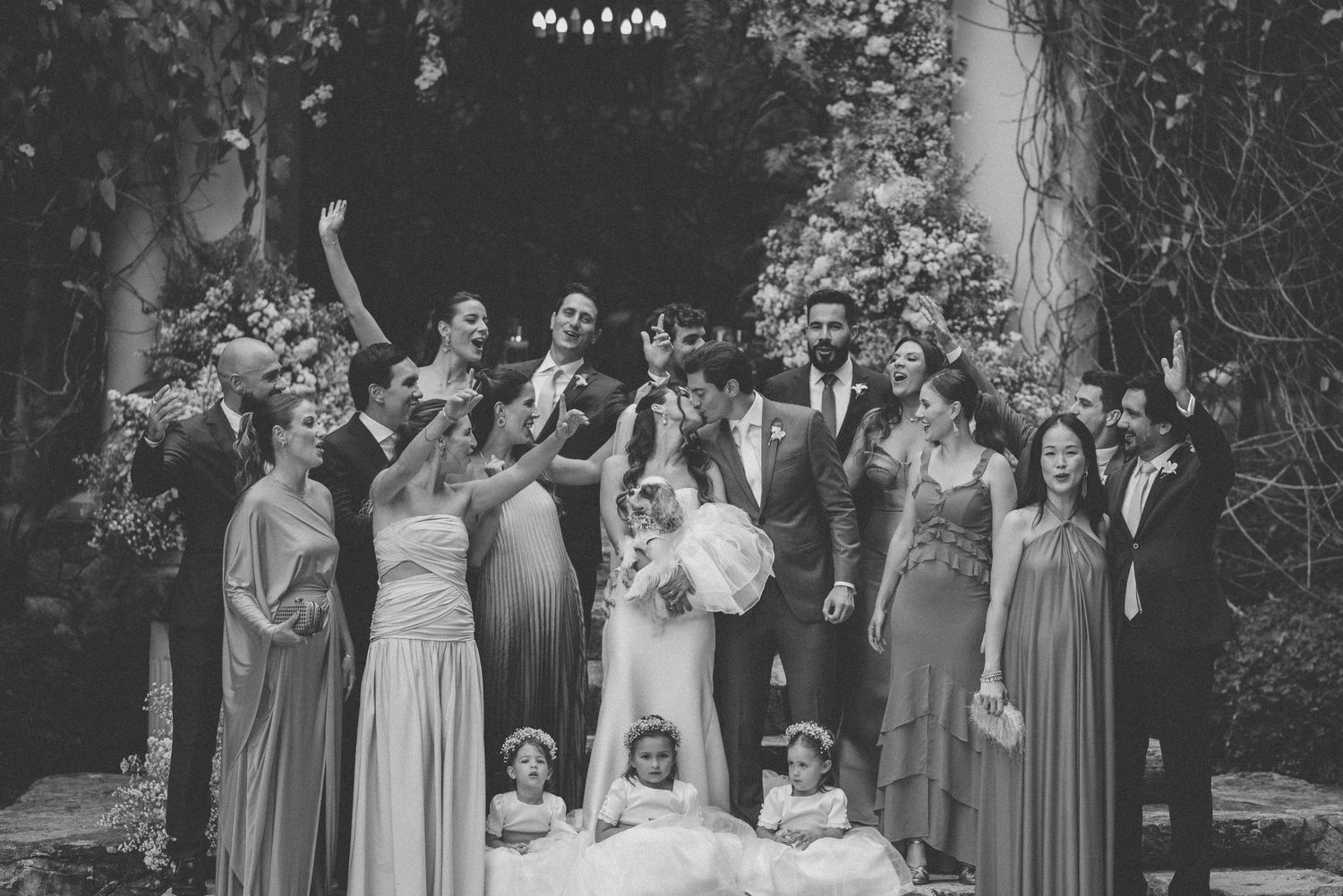 Wedding party celebrates with raised hands under garden archway in black and white photo.