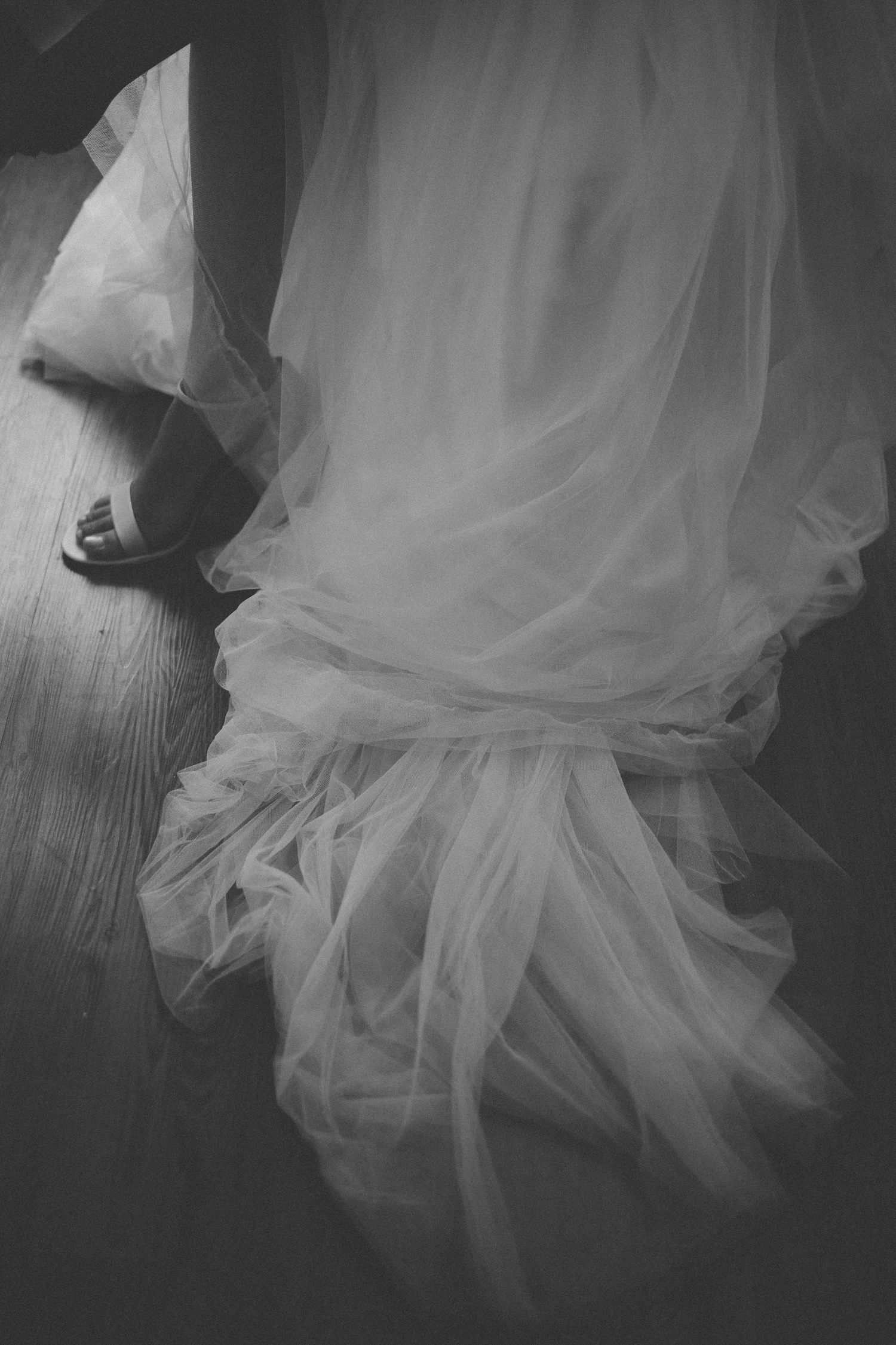 Flowing white tulle wedding dress fabric and shoe detail on wooden floor in black and white.