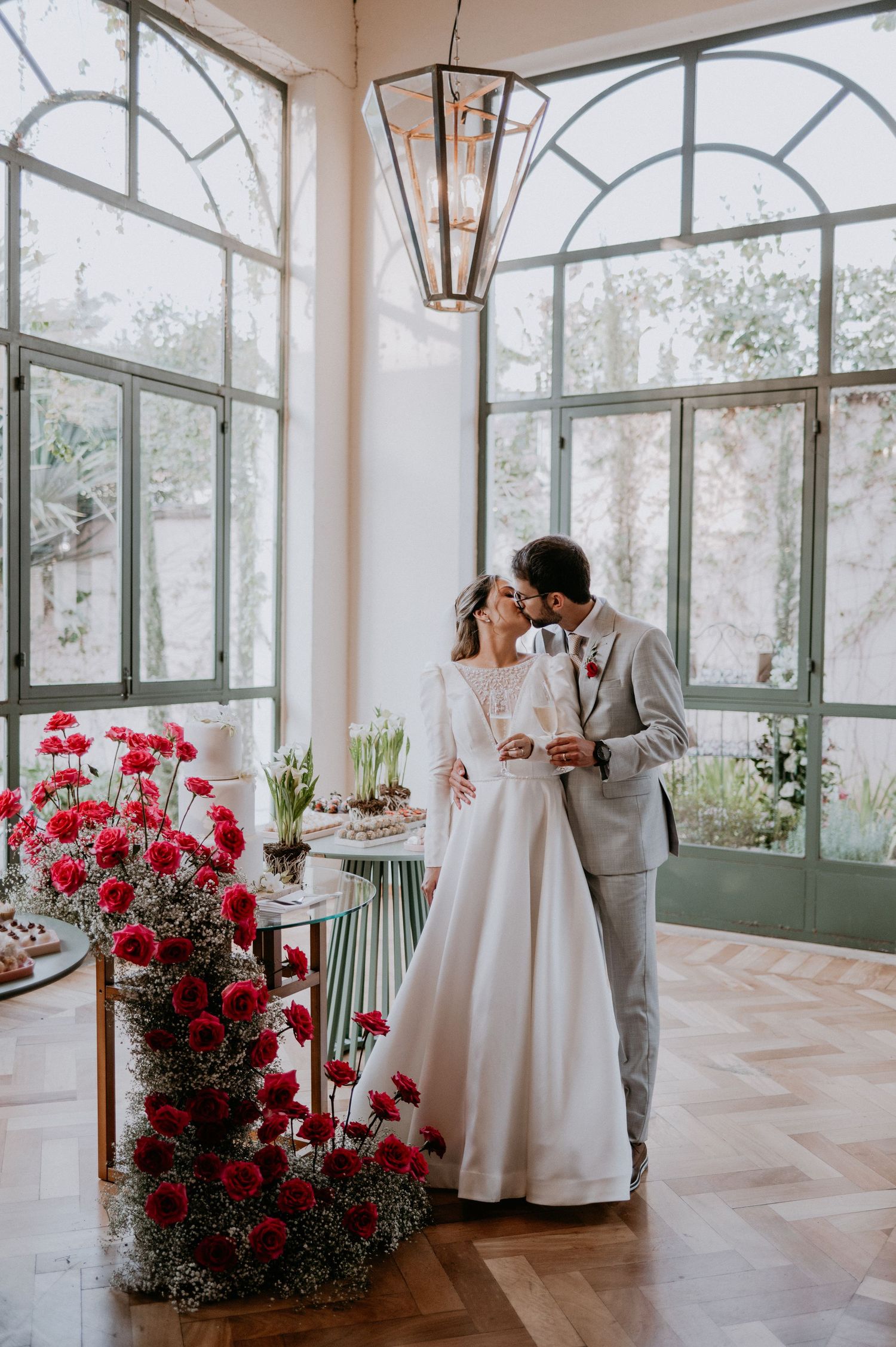 Elegant couple shares a romantic moment in a sunlit room with red floral decorations and arched windows.
