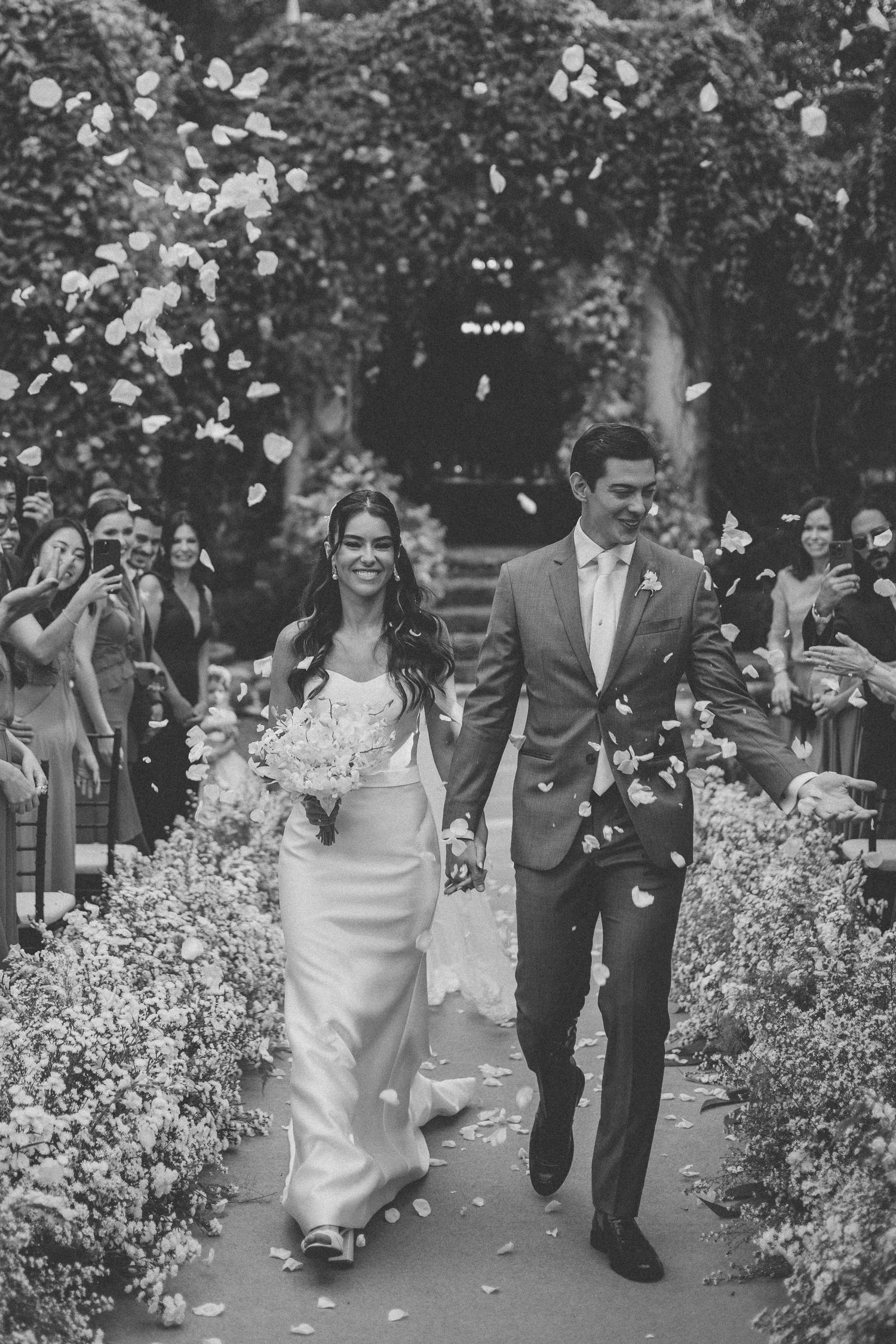 A couple walks down a garden path while guests throw flower petals in celebration.