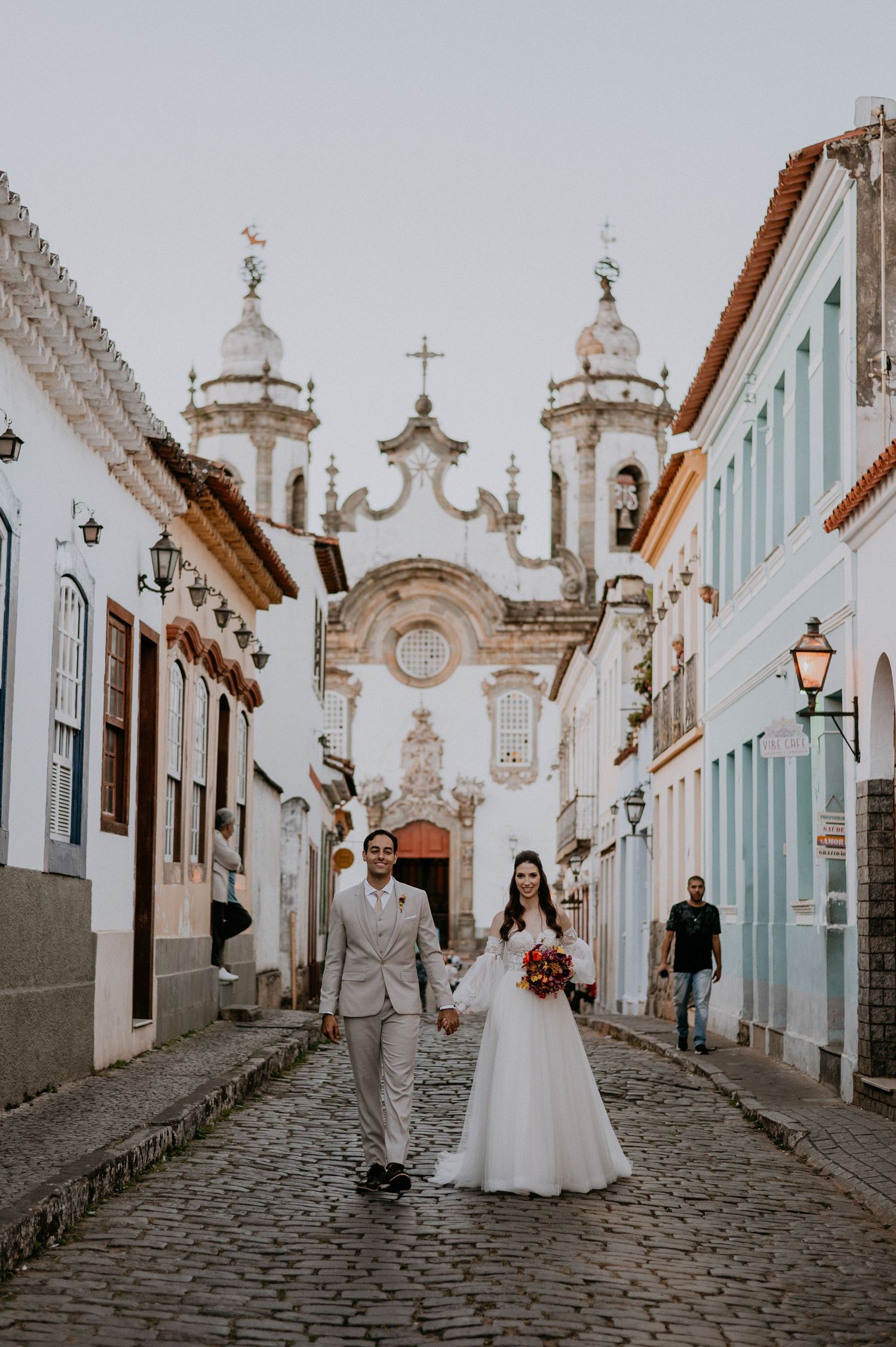 A couple walking down a cobblestone street in front of an ornate baroque church with twin towers.