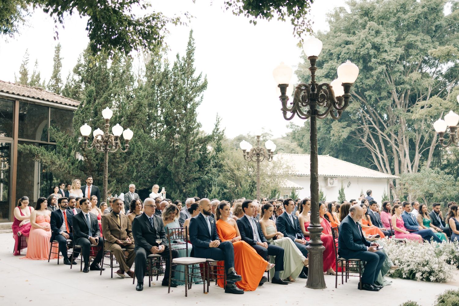 Outdoor wedding ceremony with guests seated on white chairs under trees and vintage lamp posts.
