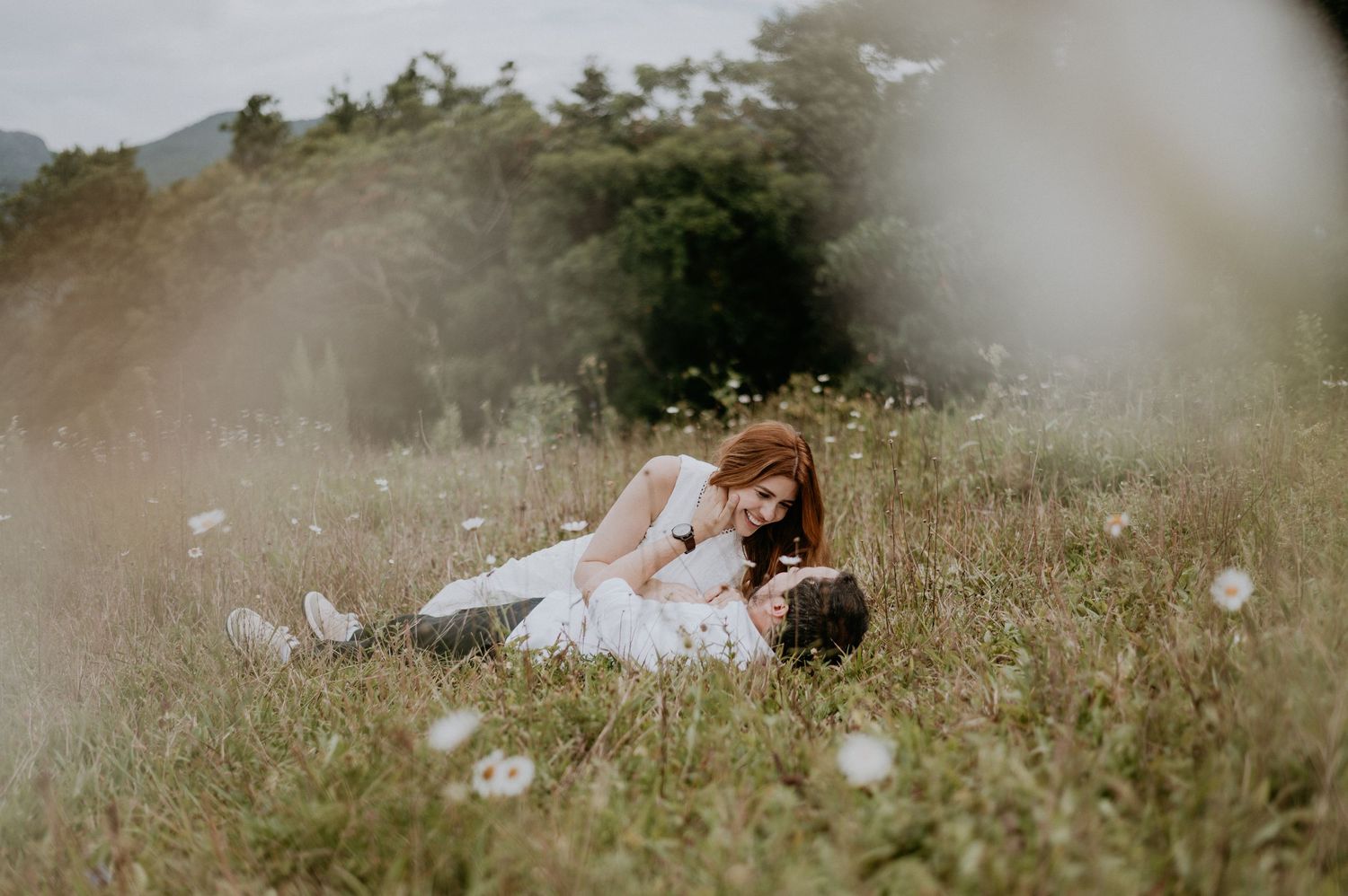 A couple in white wedding attire lies in a misty meadow surrounded by delicate wildflowers.