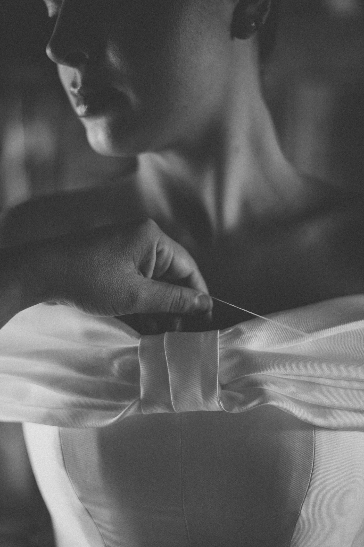 Black and white photo of a bride fastening her elegant satin wedding dress bow at the back.