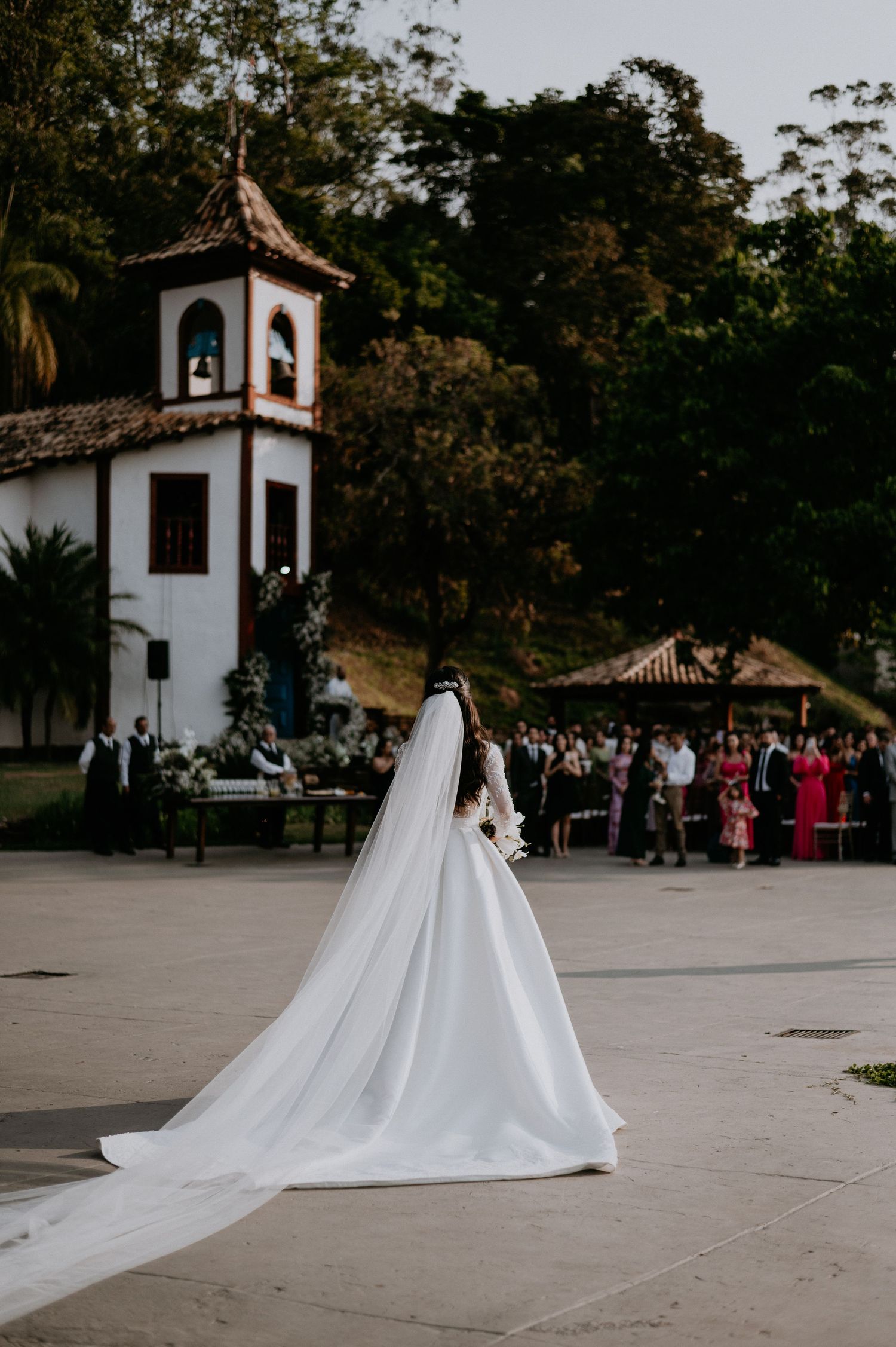 Bride in flowing white gown and veil walking toward chapel with gathered guests.