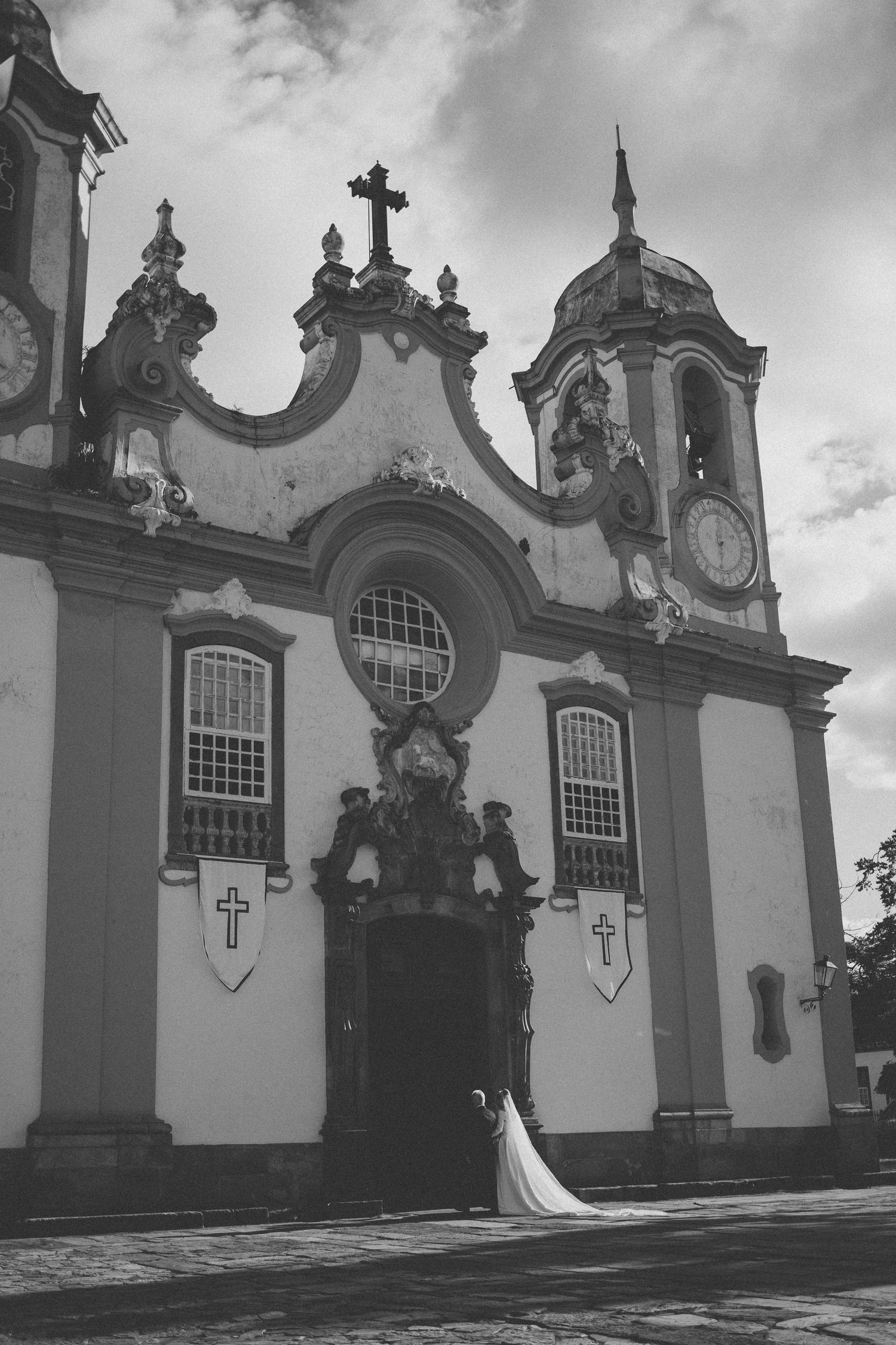 Black and white photo of a bride in a flowing gown standing before an ornate baroque church with crosses and clock tower.