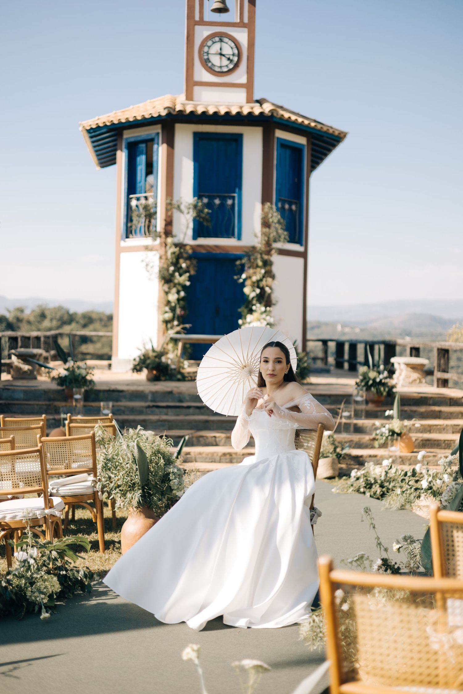 A bride in an elegant white gown poses with a parasol on a terrace featuring a distinctive clock tower and scenic backdrop.