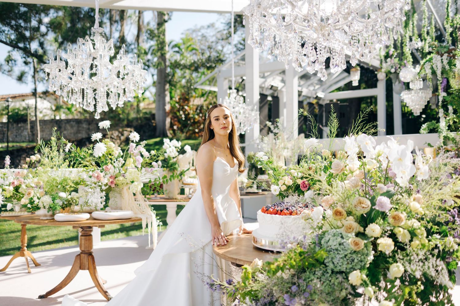 Bride in white gown surrounded by lush floral arrangements and crystal chandeliers in an elegant outdoor wedding venue.