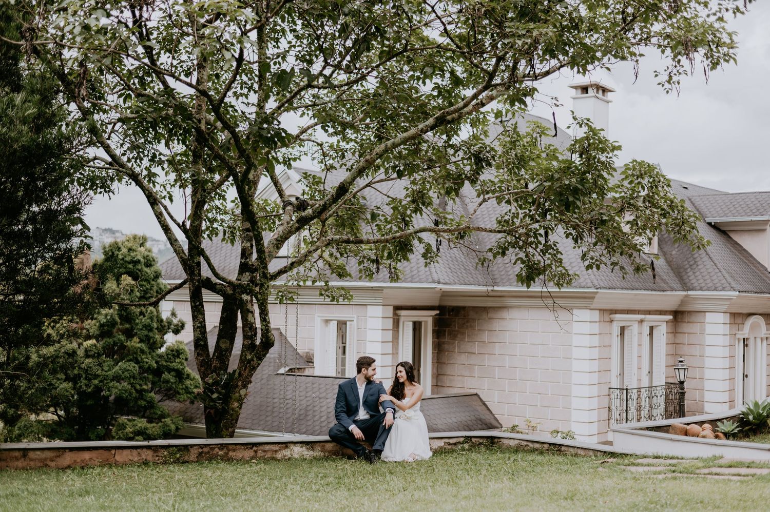 A couple sits together under a large tree on the lawn in front of an elegant white house.