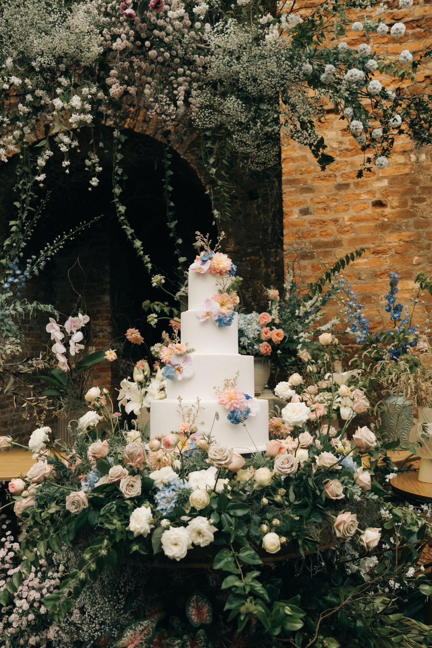 Elegant white tiered wedding cake decorated with blue and pink flowers surrounded by roses and greenery.