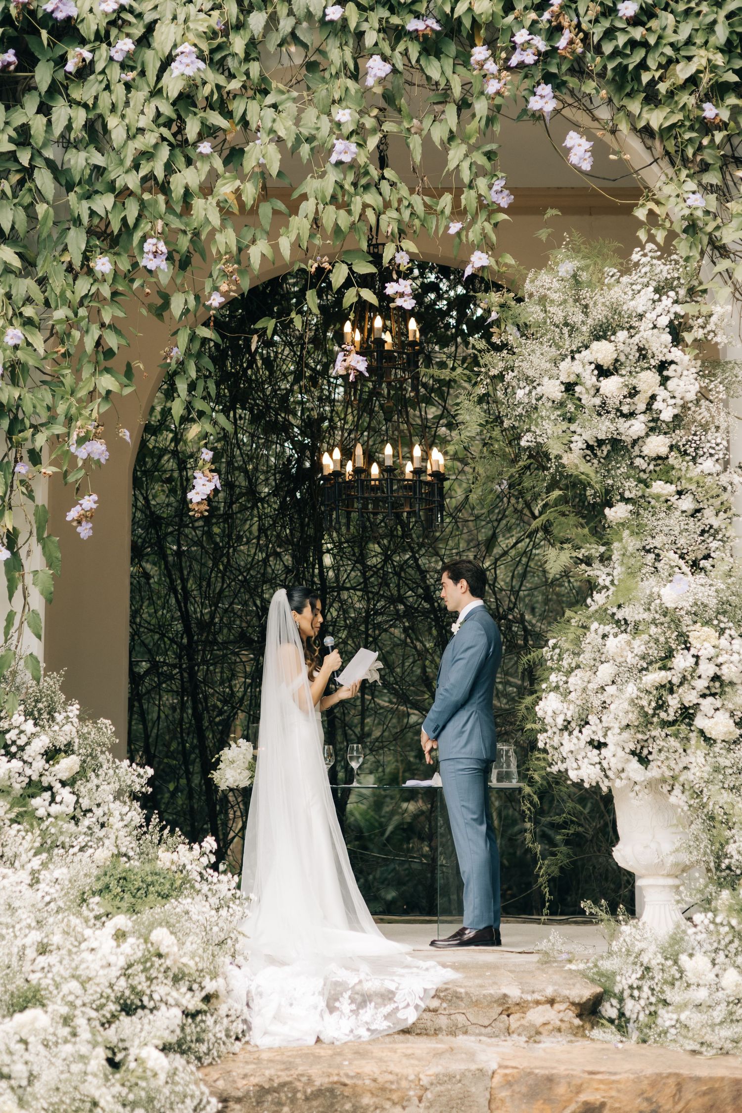 A romantic wedding ceremony takes place under a floral-adorned archway with hanging lights.