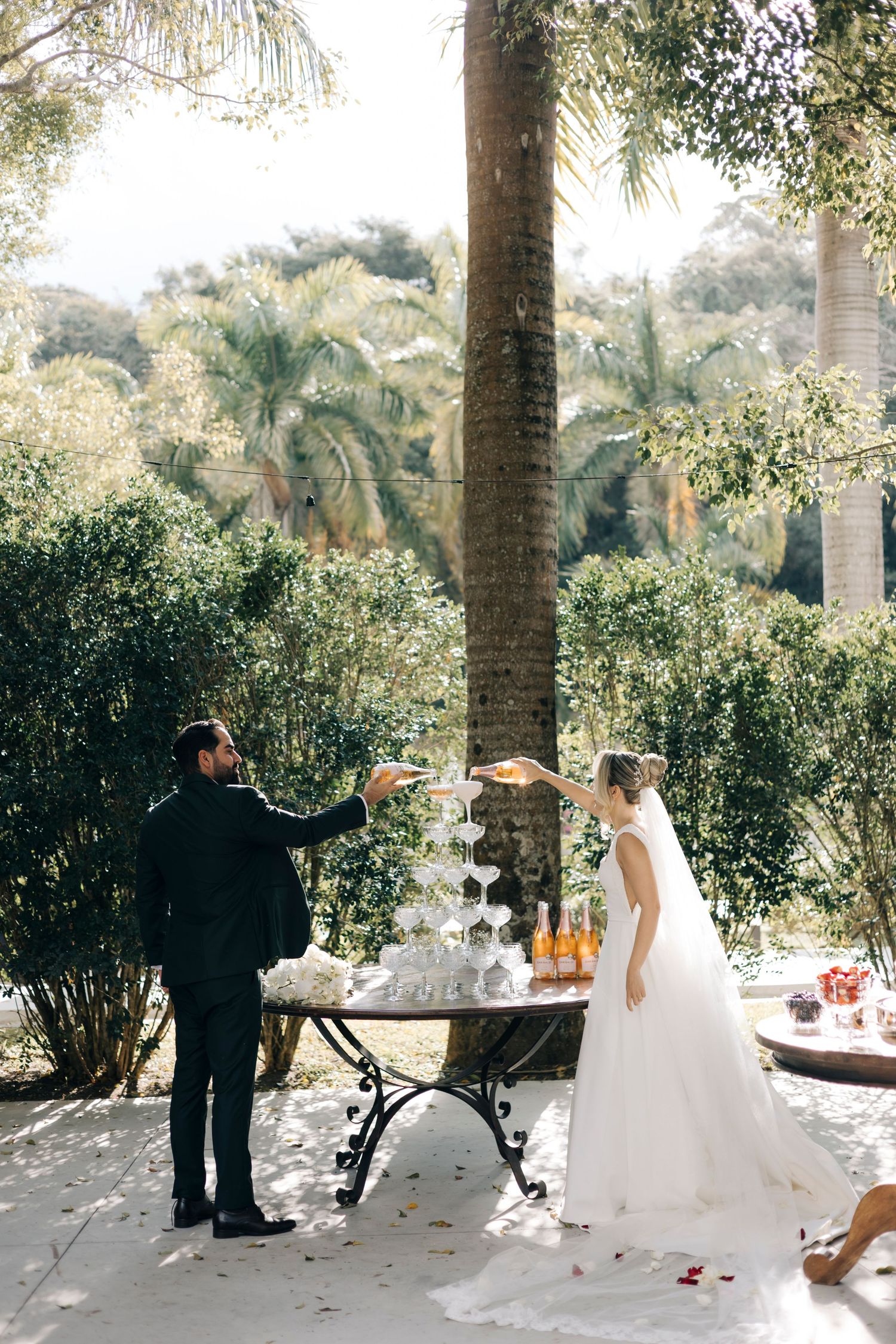 Bride and groom pouring champagne tower at outdoor wedding reception surrounded by lush tropical palm trees and greenery.