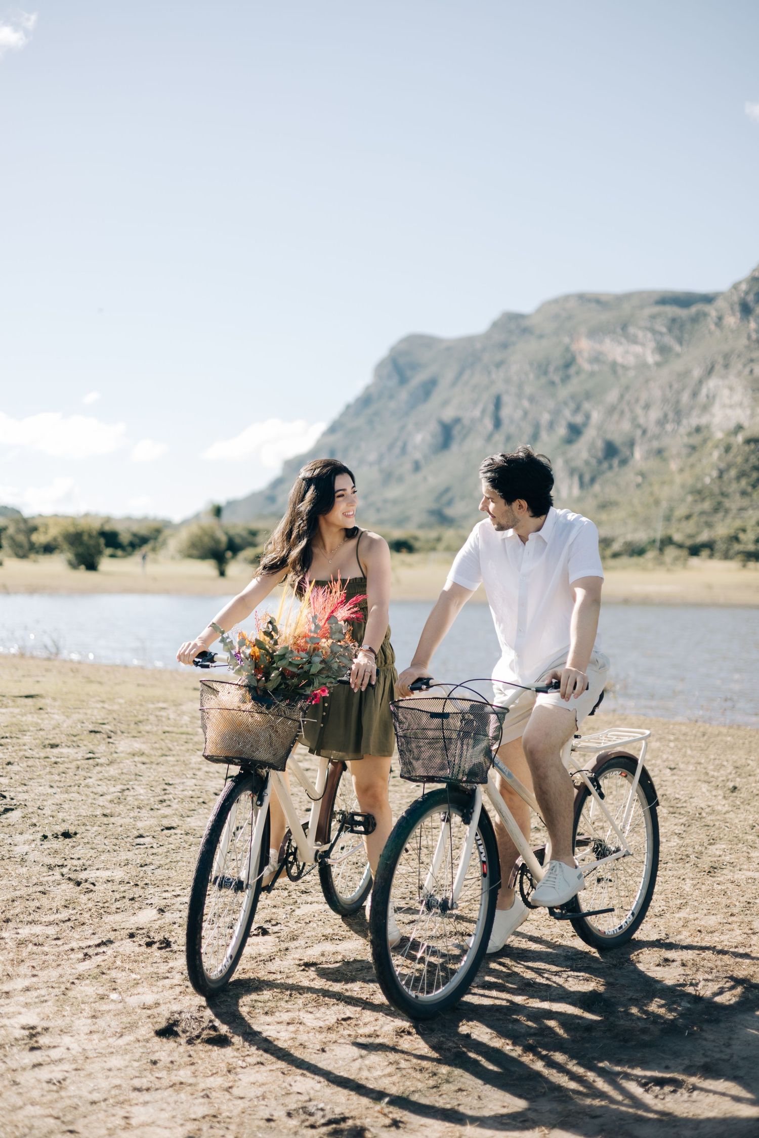 Two cyclists on vintage bikes share a romantic moment while riding along a beach with mountains in the background.